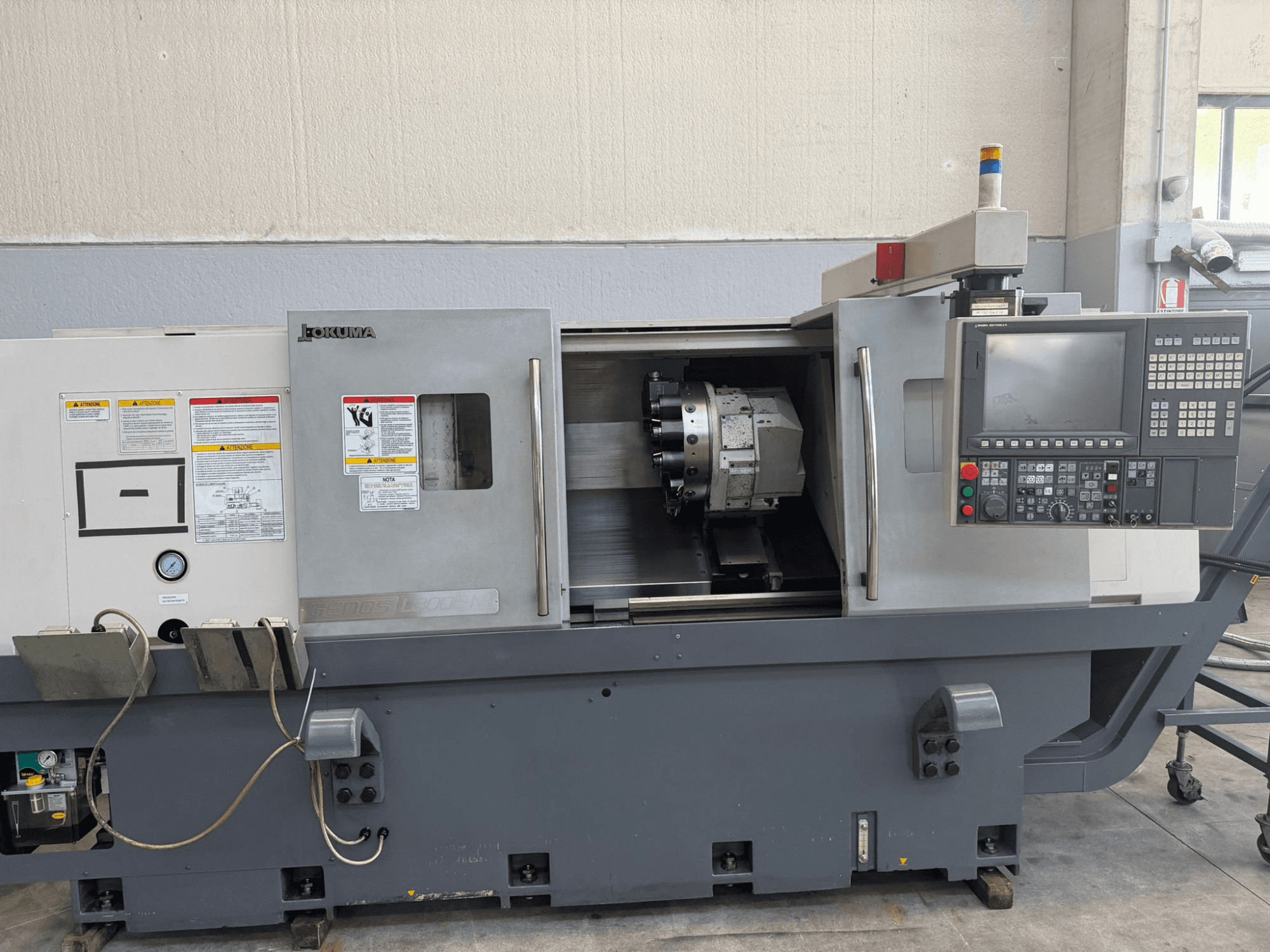 Okuma CNC virpas, priekšējais skats, kurā redzams vadības panelis, griezējinstrumentu turētājs un drošības brīdinājumi sānos.