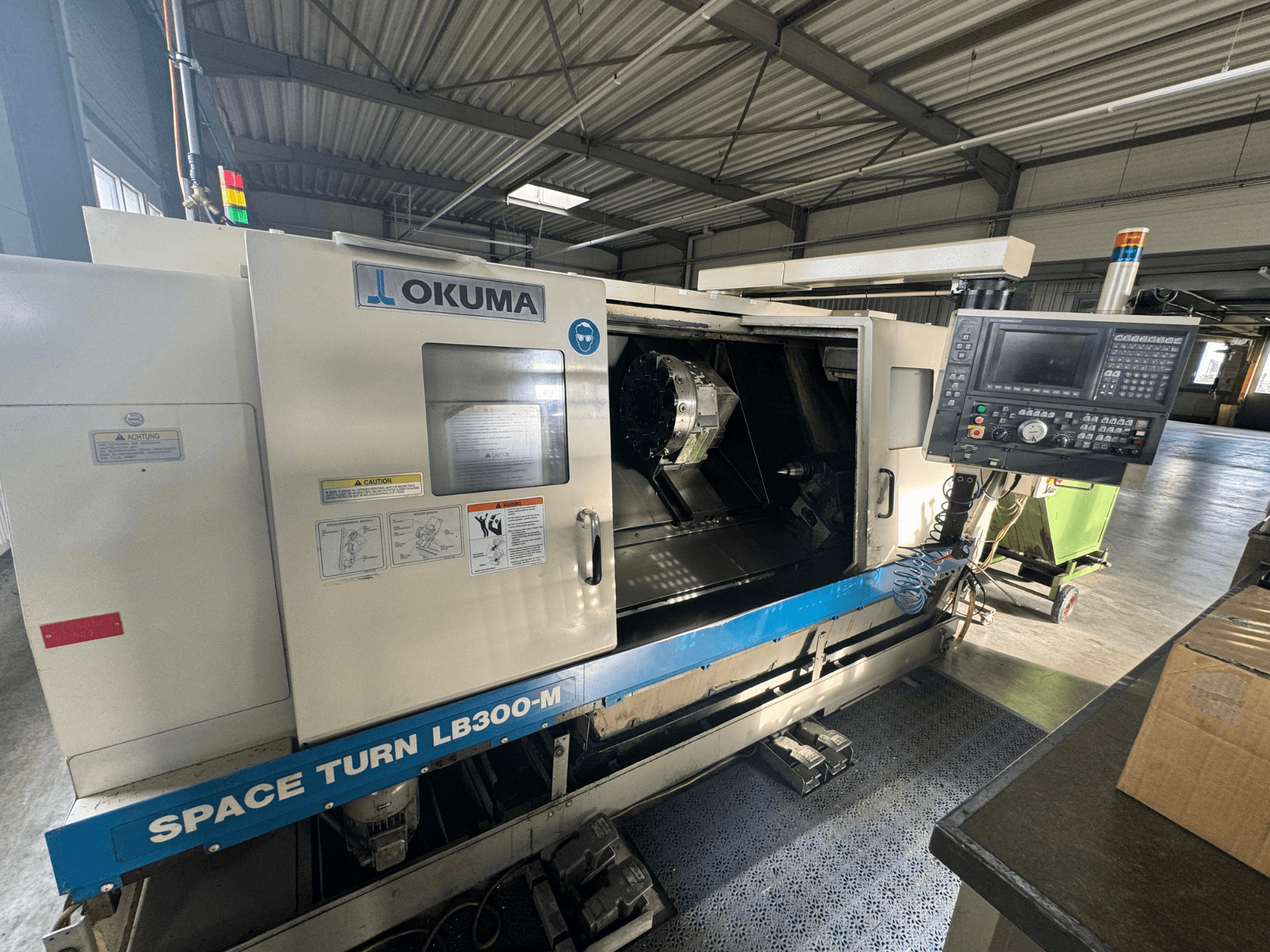 Okuma Space Turn LB300-M CNC virpa priekšpusē, ar vadības paneli un atvērtām durvīm, kas atklāj iekšējo apstrādes zonu.
