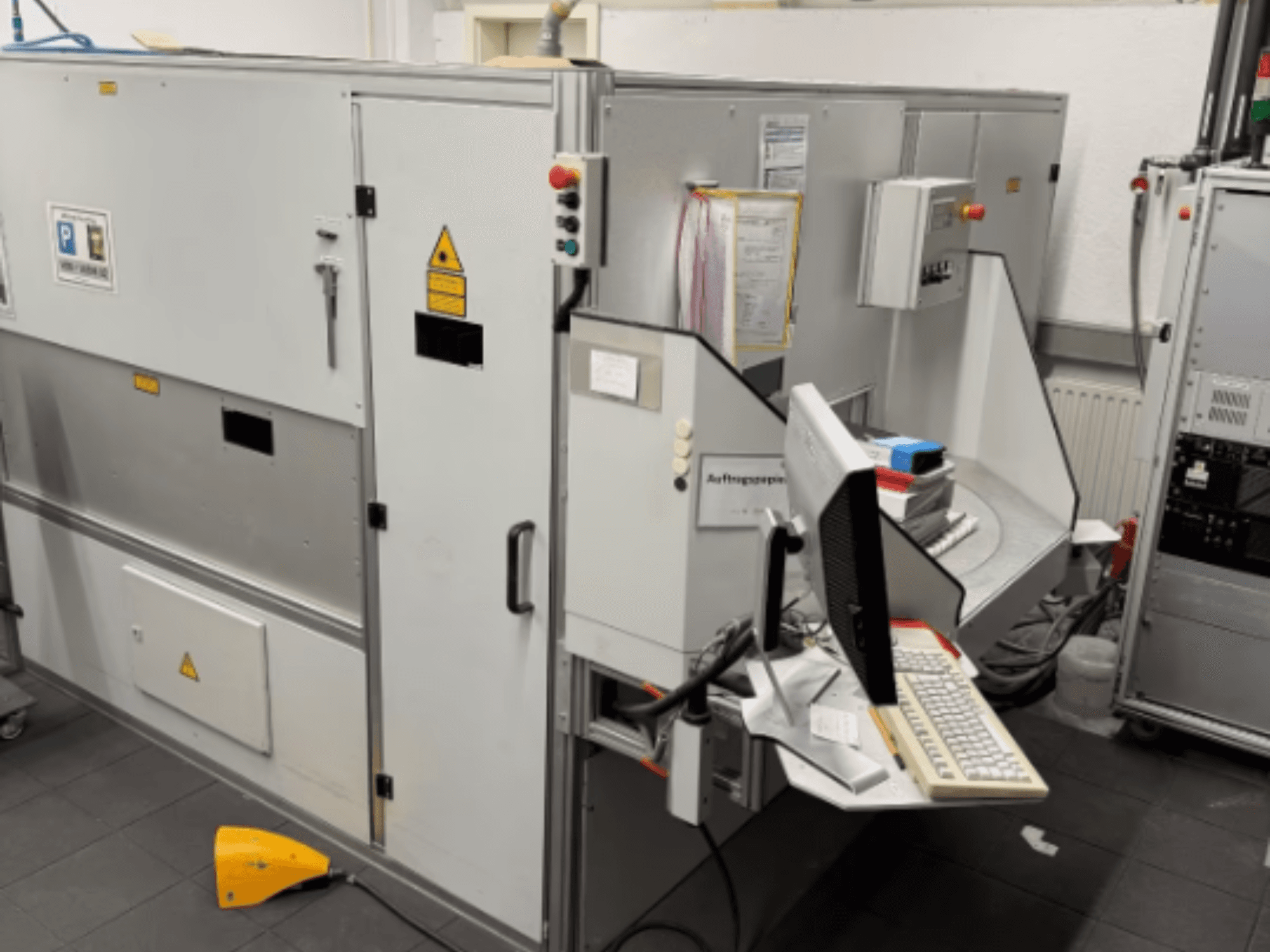 Rūpnieciskā mašīna ar datora termināli, skats no sāniem; ietver vadības pogas un drošības brīdinājuma zīmes.