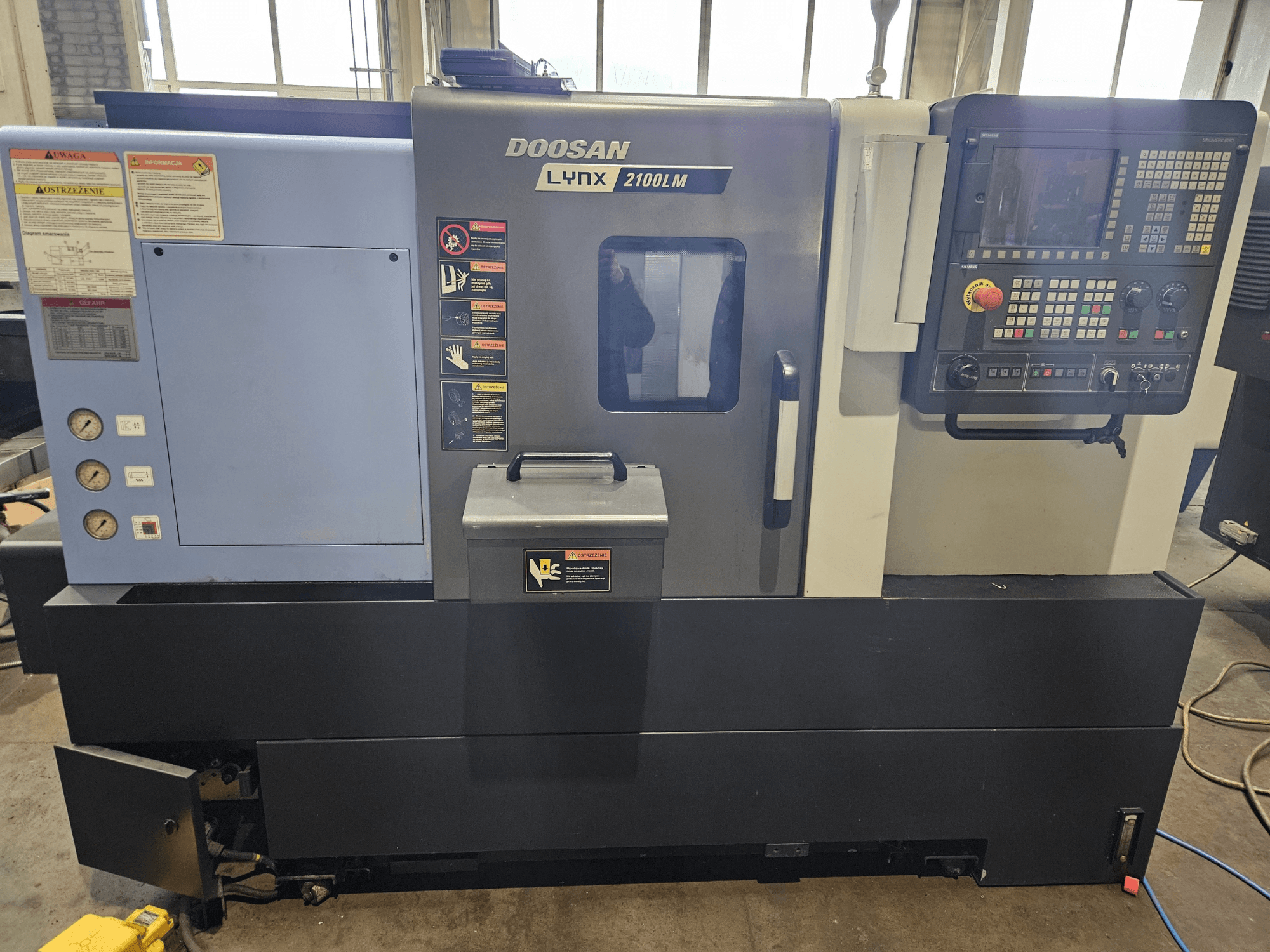 Doosan Lynx 2100LM CNC virpa, skats no priekšpuses, redzams vadības panelis, gaisa spiediena mērinstrumenti un drošības marķējums.