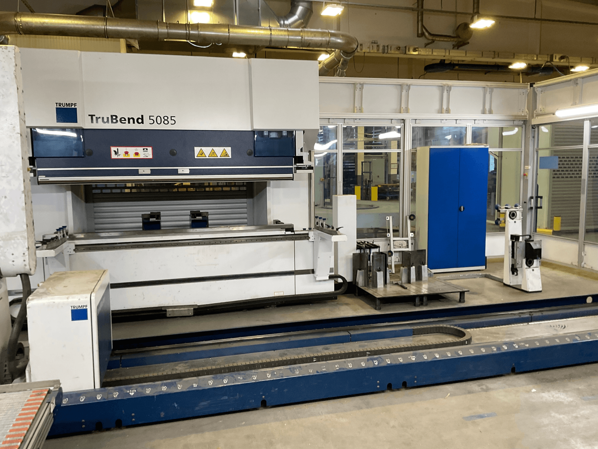 TRUMPF TruBend 5085, skats no sāniem uz lielu metāla liešanas iekārtu ar vadības paneli un dažādiem instrumentiem blakus.