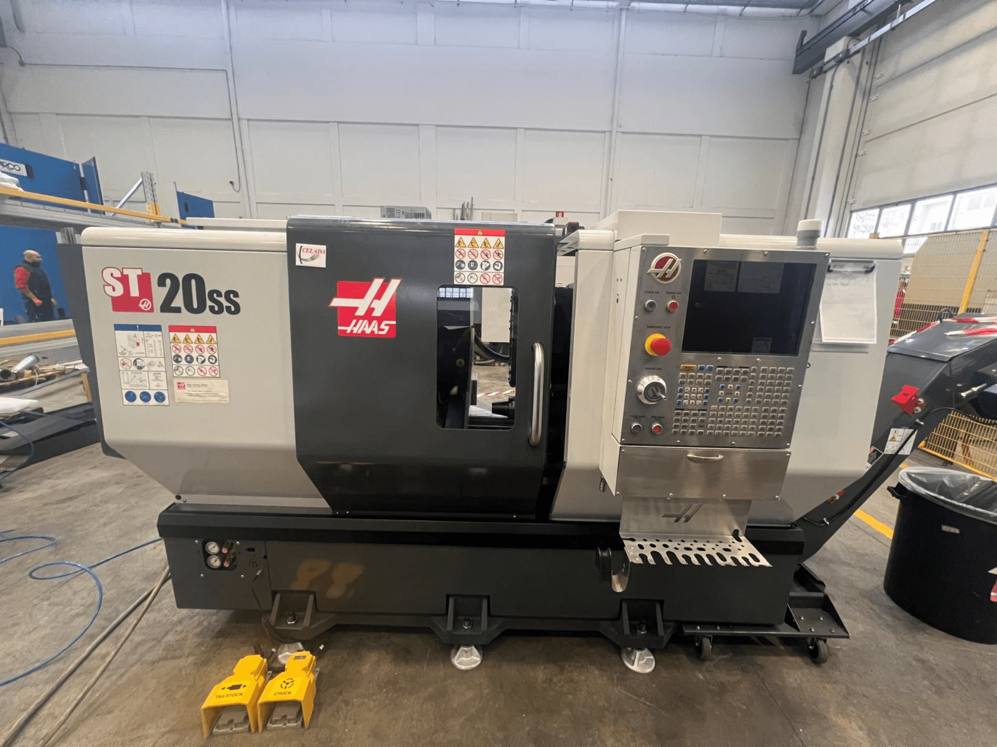 Haas ST 20SS CNC virpa sānskatā, ar vadības paneli, drošības marķējumiem un kājstarpēm uz grīdas.