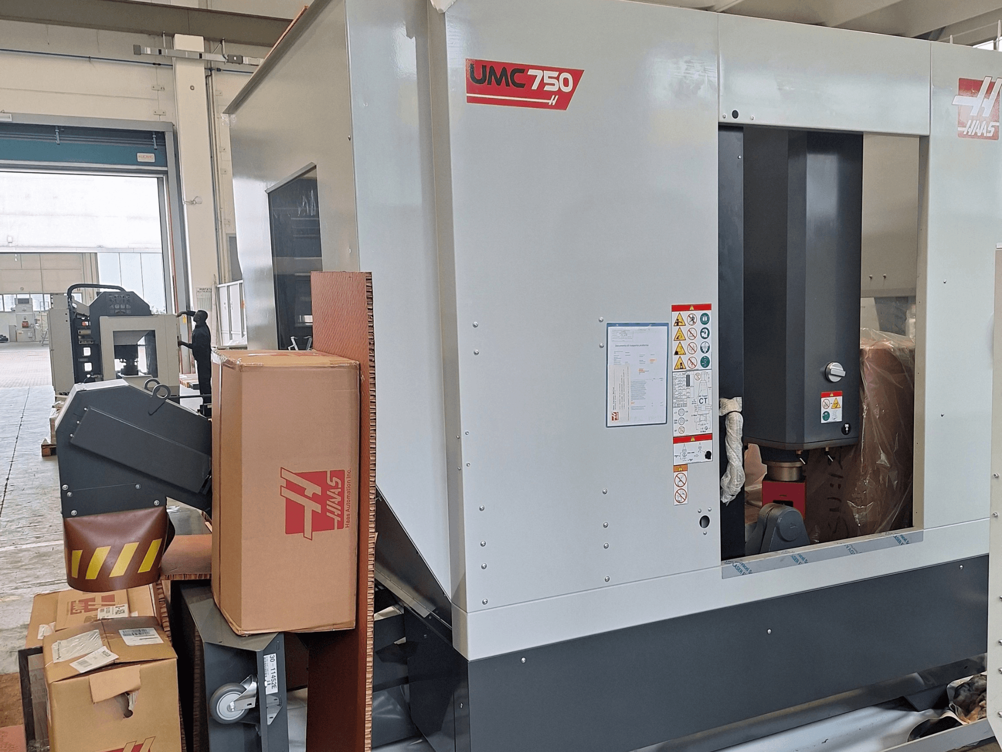 HAAS UMC-750 universālais apstrādes centrs, skats no sāniem, kurā redzams vadības panelis un instrumentu glabāšanas vieta ar brīdinājuma uzlīmēm un iepakojumu.