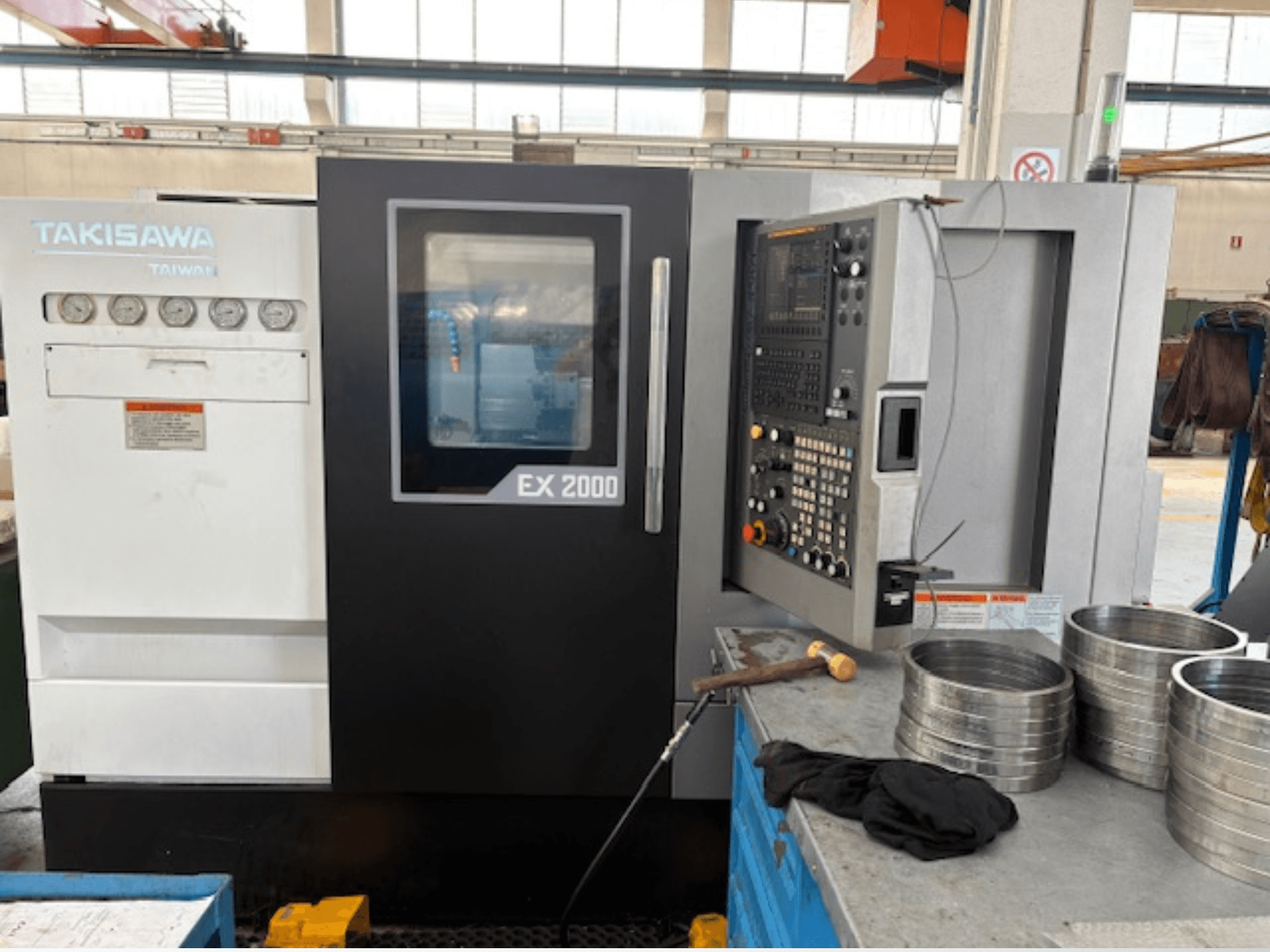 Takisawa EX 2000 CNC darbagalds, skats no priekšpuses, ar vadības paneli, mērinstrumentiem un metāla detaļām uz darbnīcas galda.