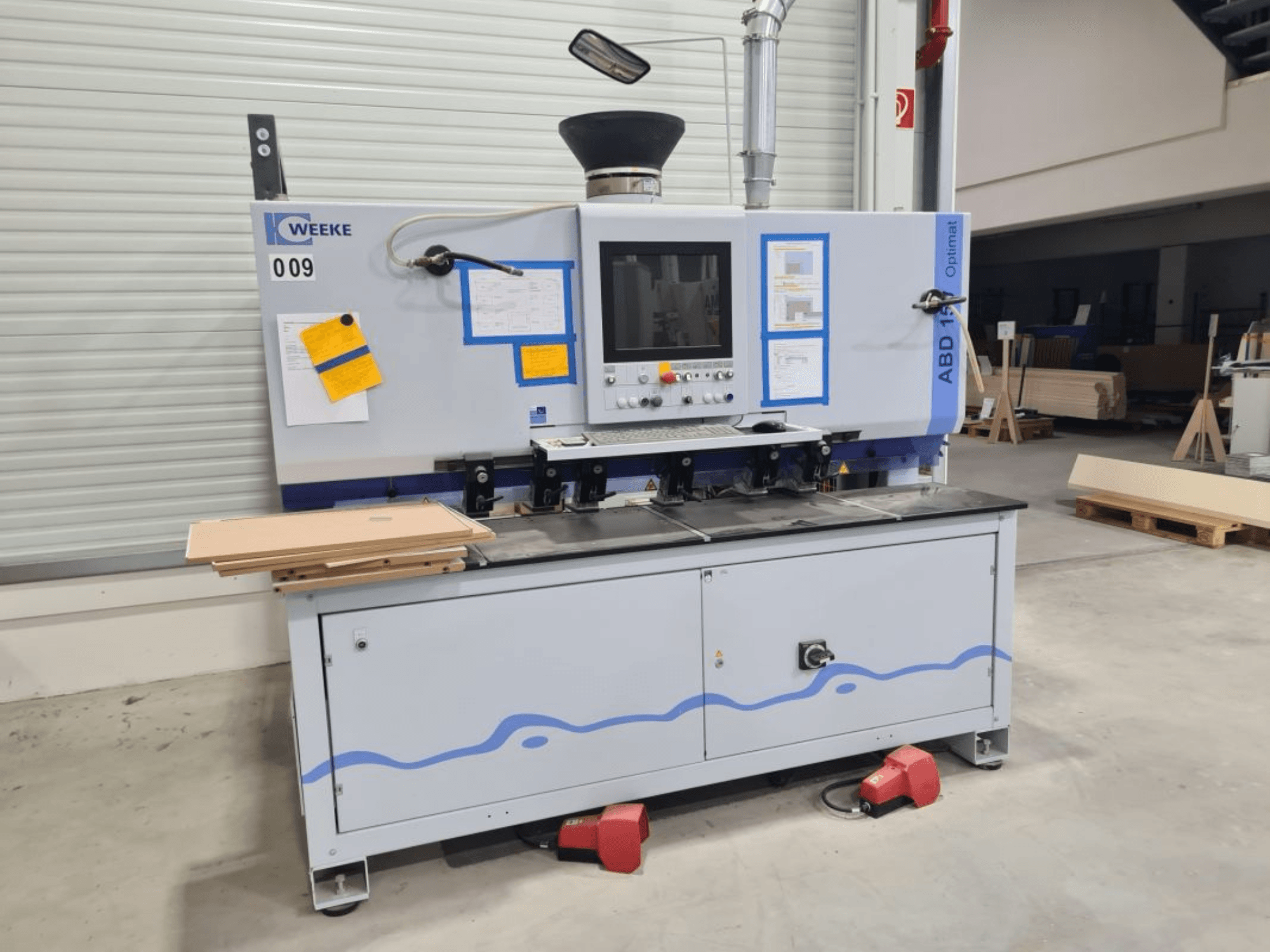 HOMAG WEEKE OPTIMAT ABD150 daudzvārpstu urbjmašīna, skats no priekšpuses, redzamas vadības ierīces un sakrauti koka paneļi.