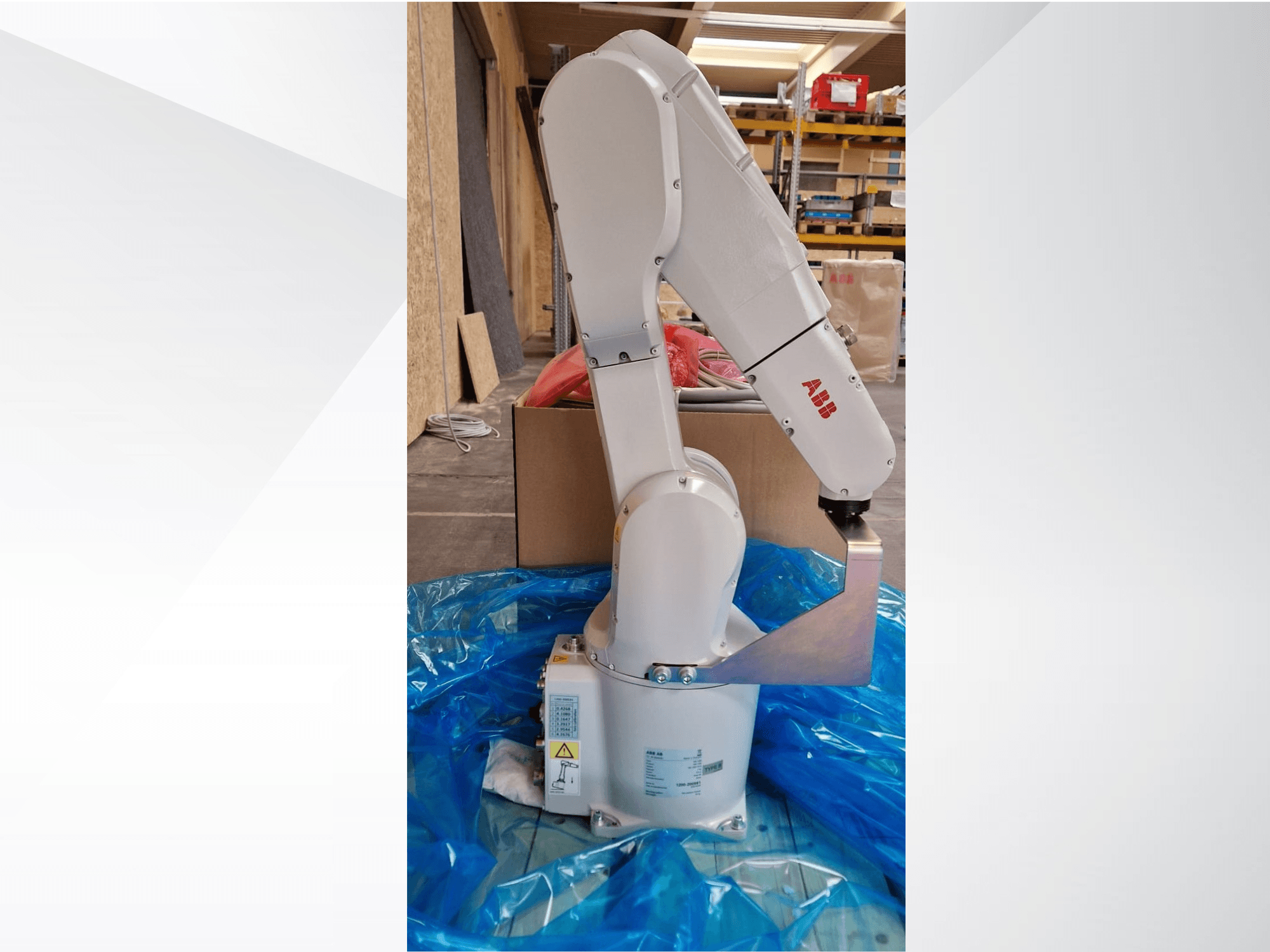 ABB robotizētās rokas modelis, skats no sāniem uz zila brezenta, ar redzamām tehniskajām uzlīmēm, kurā redzami locītavu savienojumi un metāla gala efektofors.