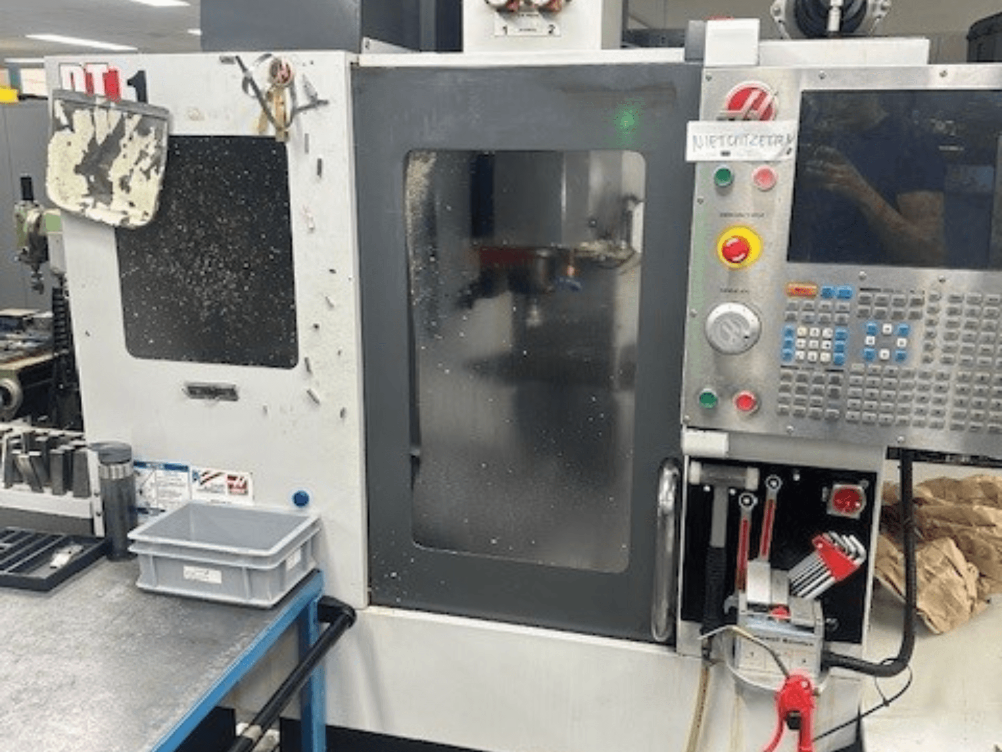 Haas DT-1 vertikālās apstrādes centrs, priekšējais skats ar vadības paneli, redzamiem netīrumiem un instrumentu paliktni uz darbgalda.