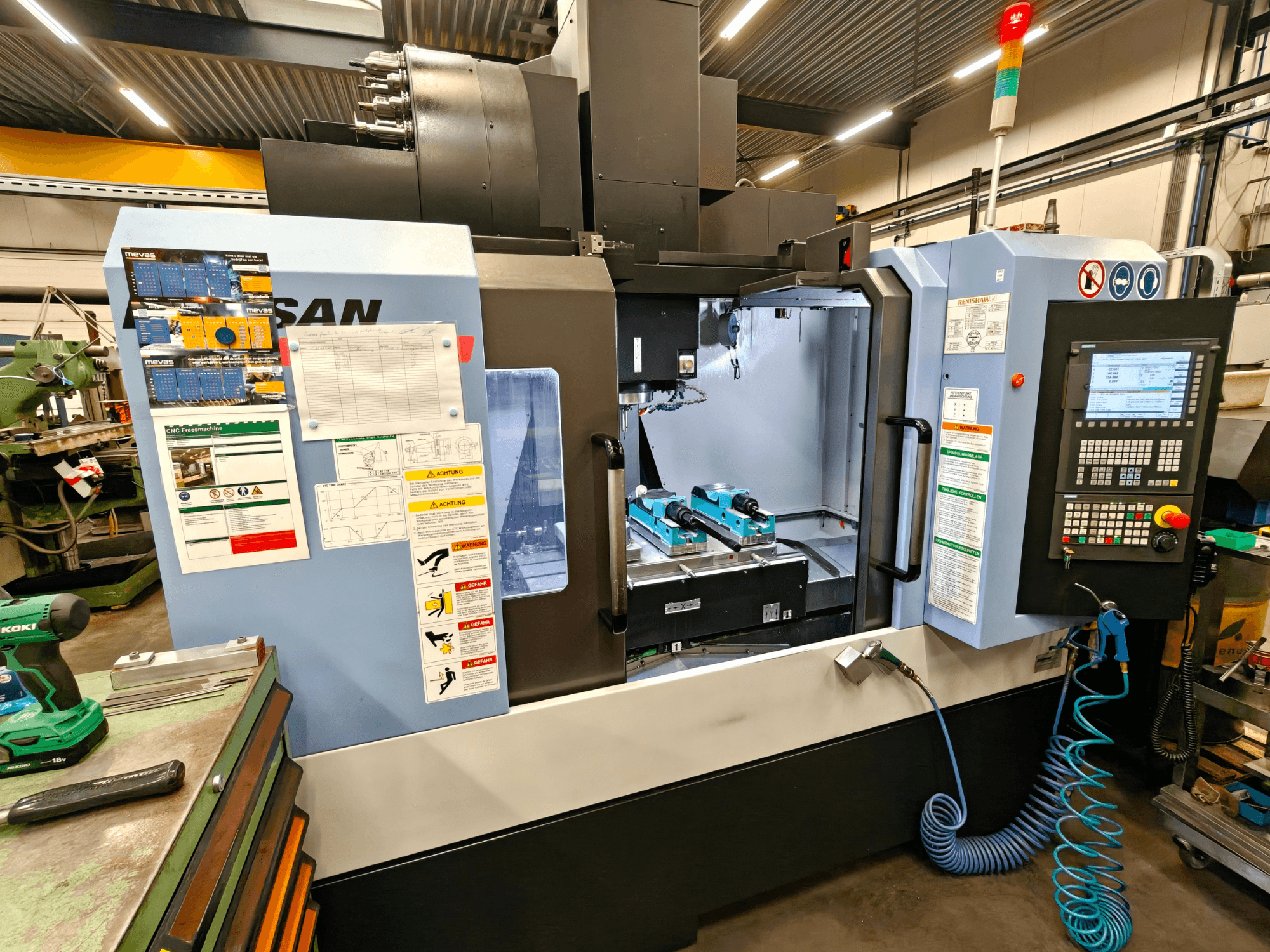 DOOSAN DNM 400 II vertikālais apstrādes centrs darbnīcā, ar vadības ierīcēm, instrumentu turētājiem un drošības zīmēm, skats no priekšpuses.