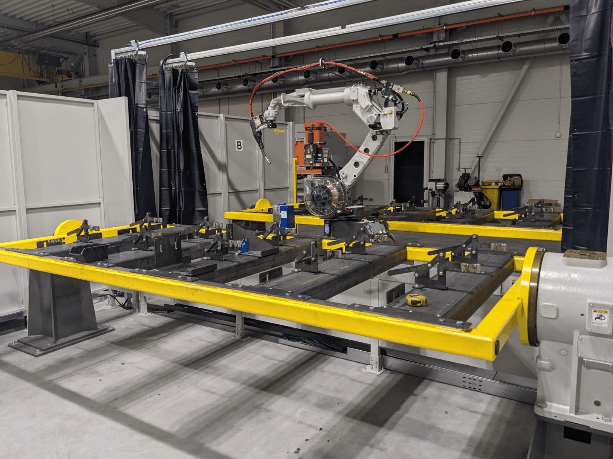 PANASONIC TAWERS TM-2000WG3 robota rokas skats no priekšpuses, kas novietota virs dzeltena darba galda ar montāžai paredzētām metāla detaļām.