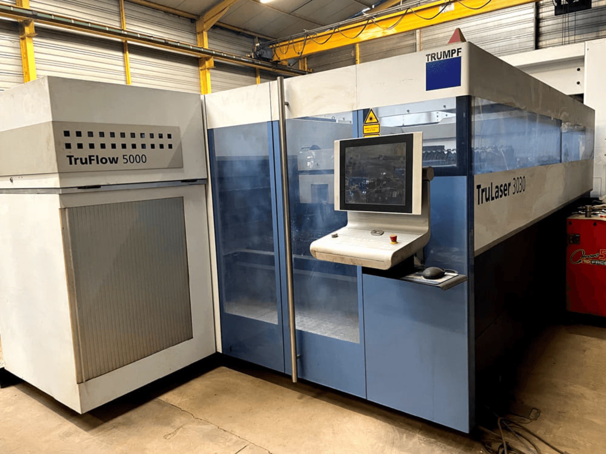 TRUMPF TruLaser 3030 CO₂ lāzergriešanas iekārta, skats no priekšpuses, kurā redzams vadības panelis un blakus novietotais TruFlow 5000 bloks.