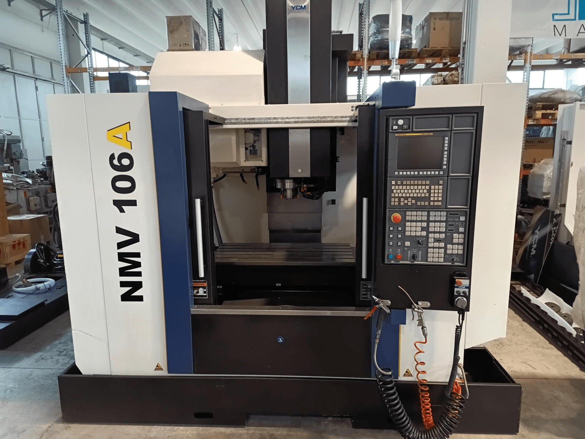 YCM NMV 106A apstrādes centrs, skats no priekšpuses, ar vadības paneli ar pogām un dzesēšanas šķidruma šļūtenēm.