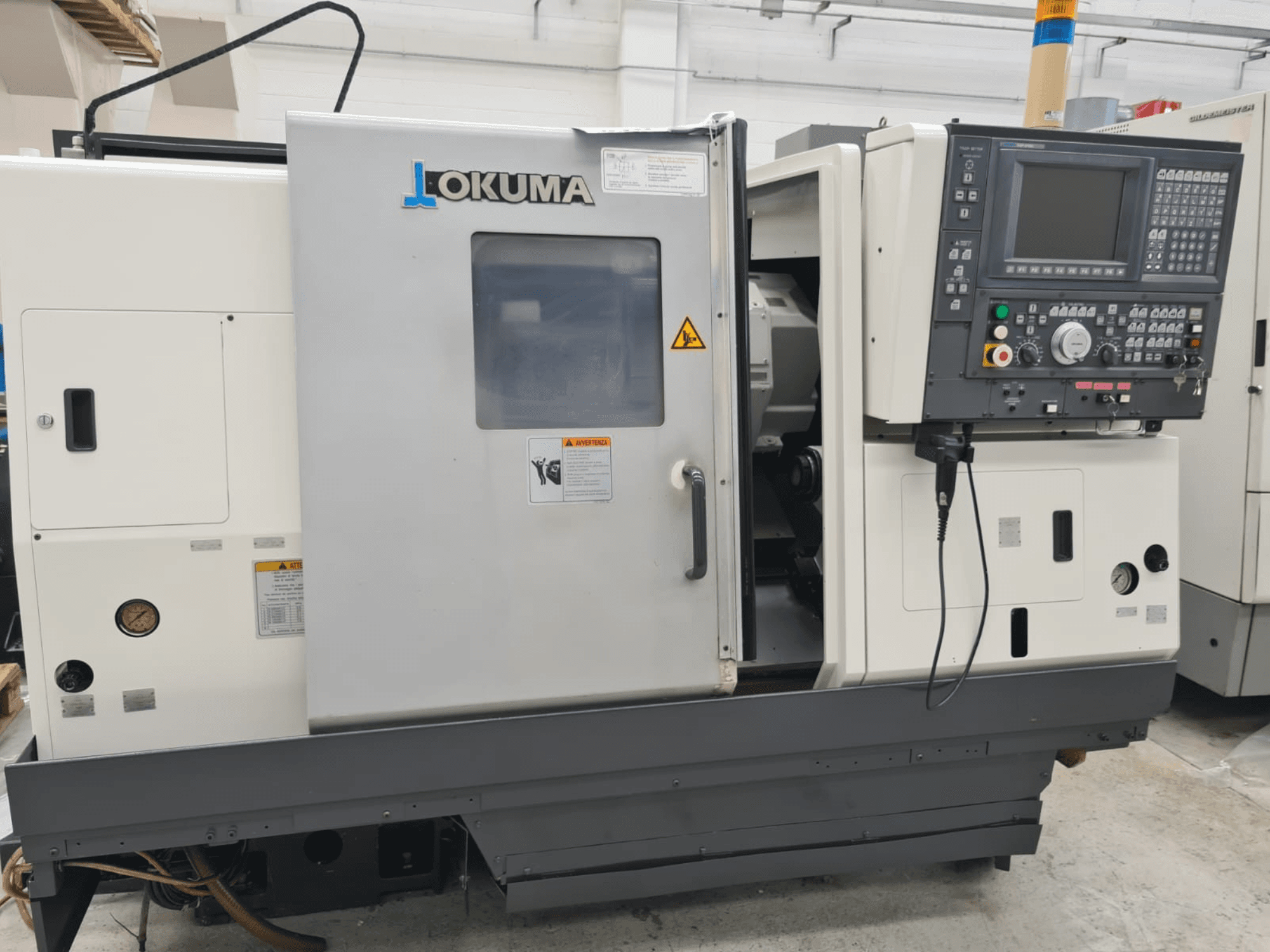 Okuma LB 300M X 500 horizontālās virpošanas darbgalds sānu skatā, kurā redzams vadības panelis, piekļuve vārpstai un drošības brīdinājumi.
