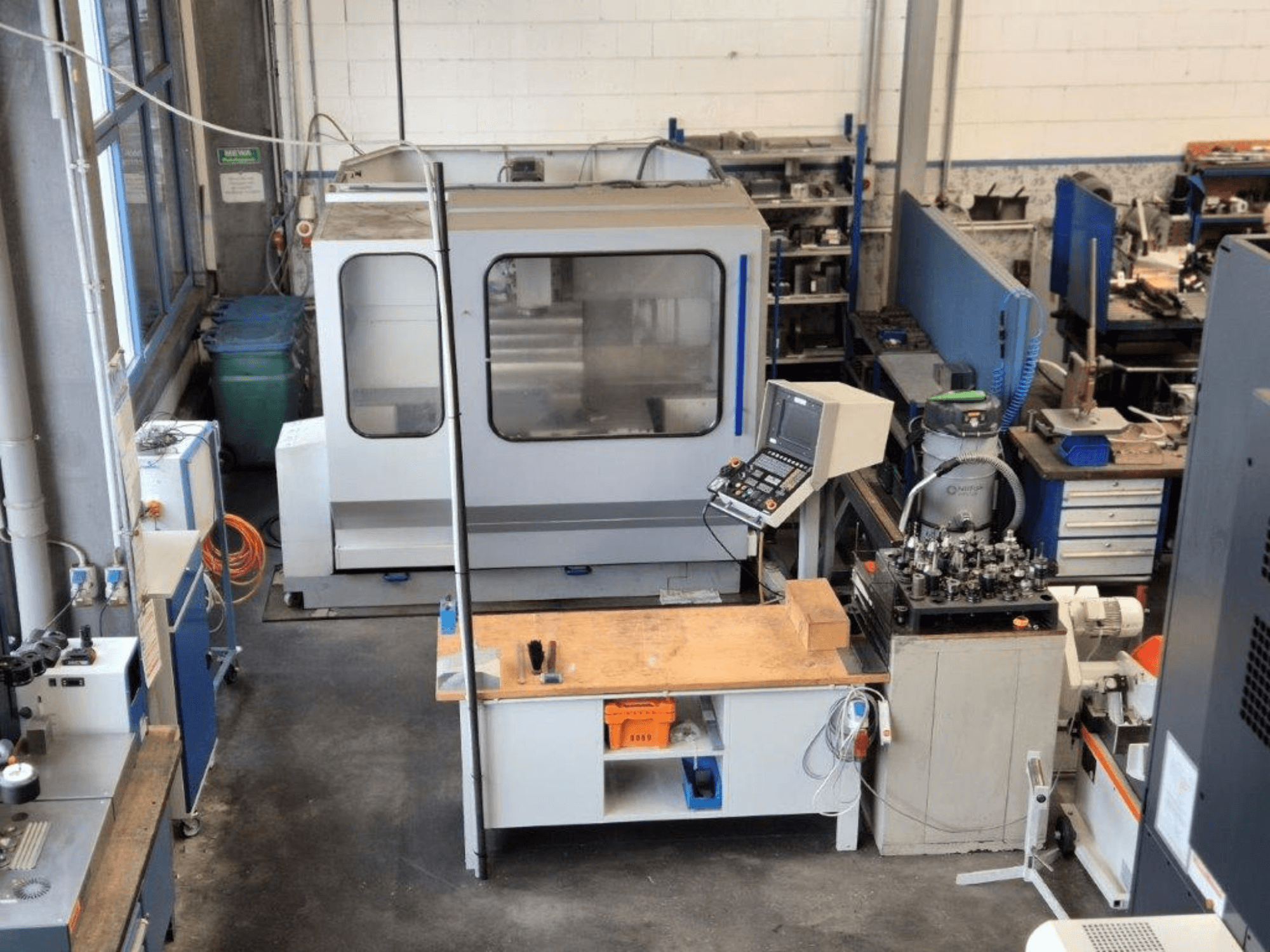 CNC frēzmašīna darbnīcā, skats no priekšpuses, fonā redzams vadības panelis, darba virsma un instrumenti.