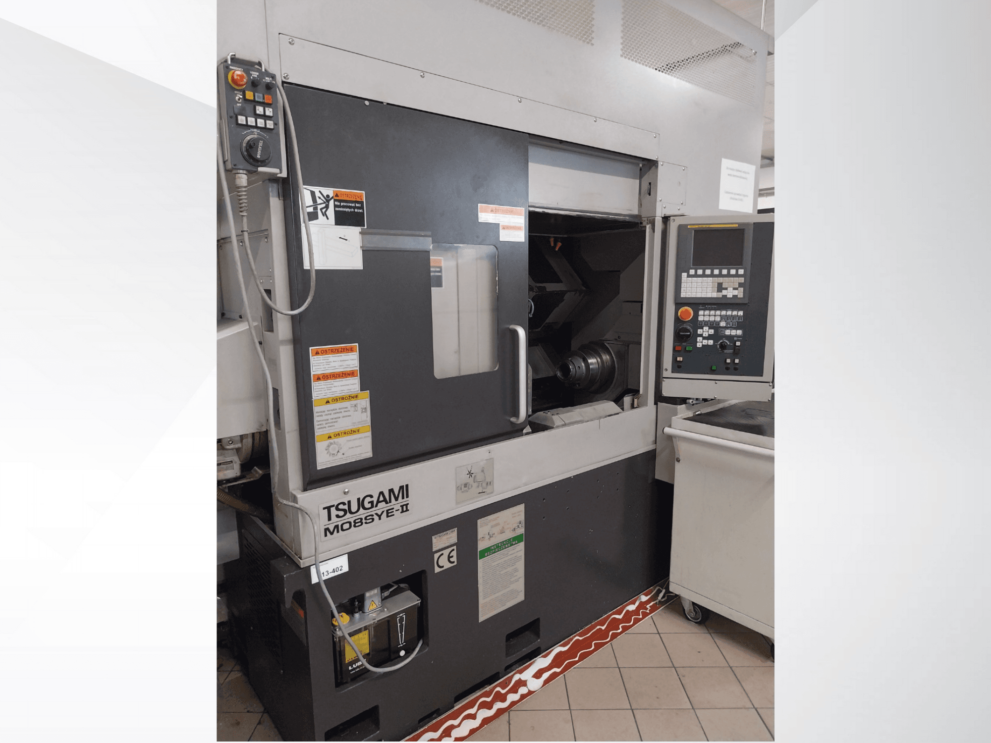 Tsugami M08SY-II CNC virpa, priekšējais skats, kurā redzams vadības panelis, vārpsta un iekraušanas zona. Ietver drošības paziņojumus un zīmola etiķeti.