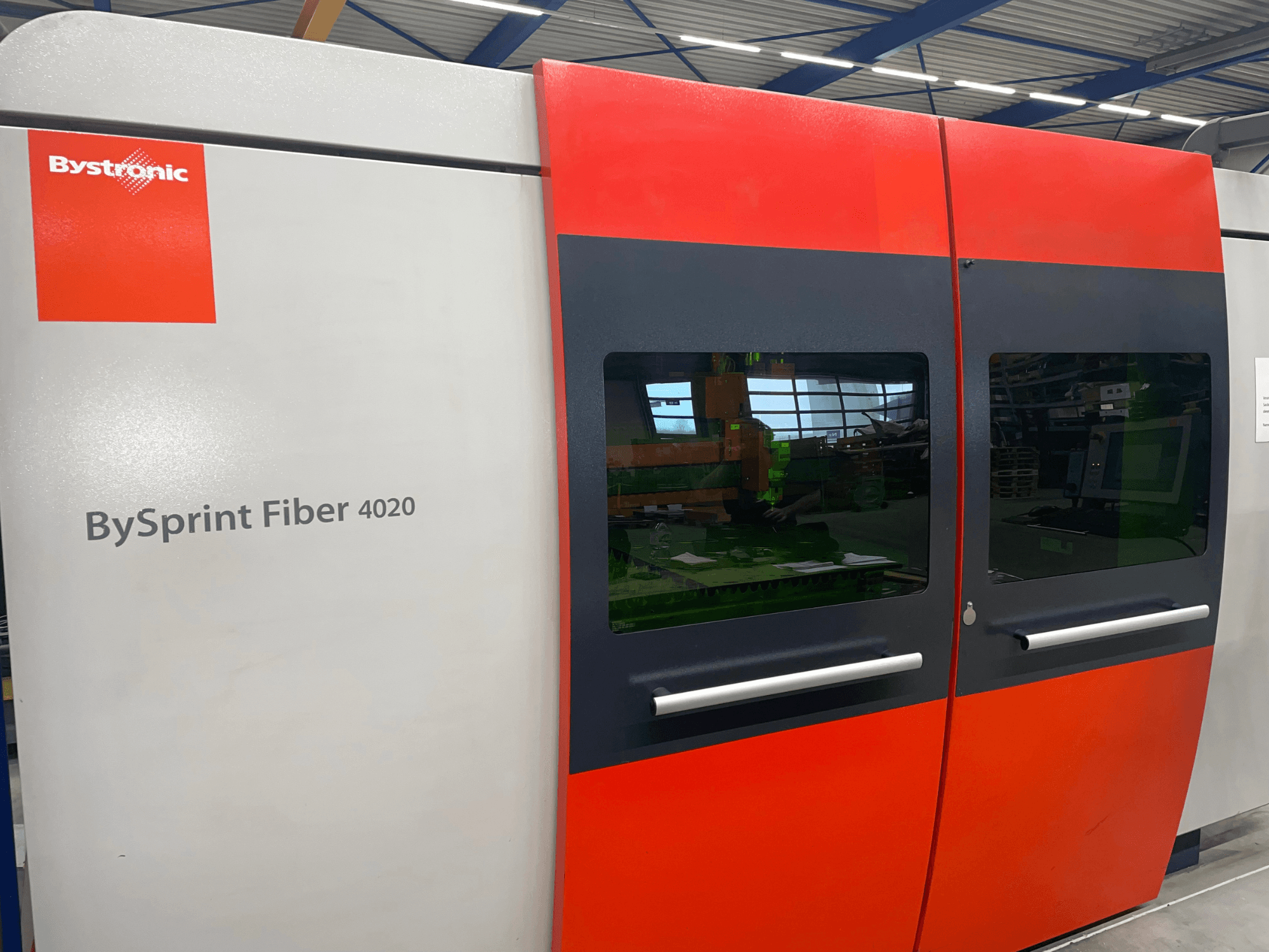 Bystronic BySprint Fiber 4020 no priekšpuses, ar pelēku un sarkanu paneļa dizainu ar iekšpusē redzamiem lieliem logiem un vadības saskarni.