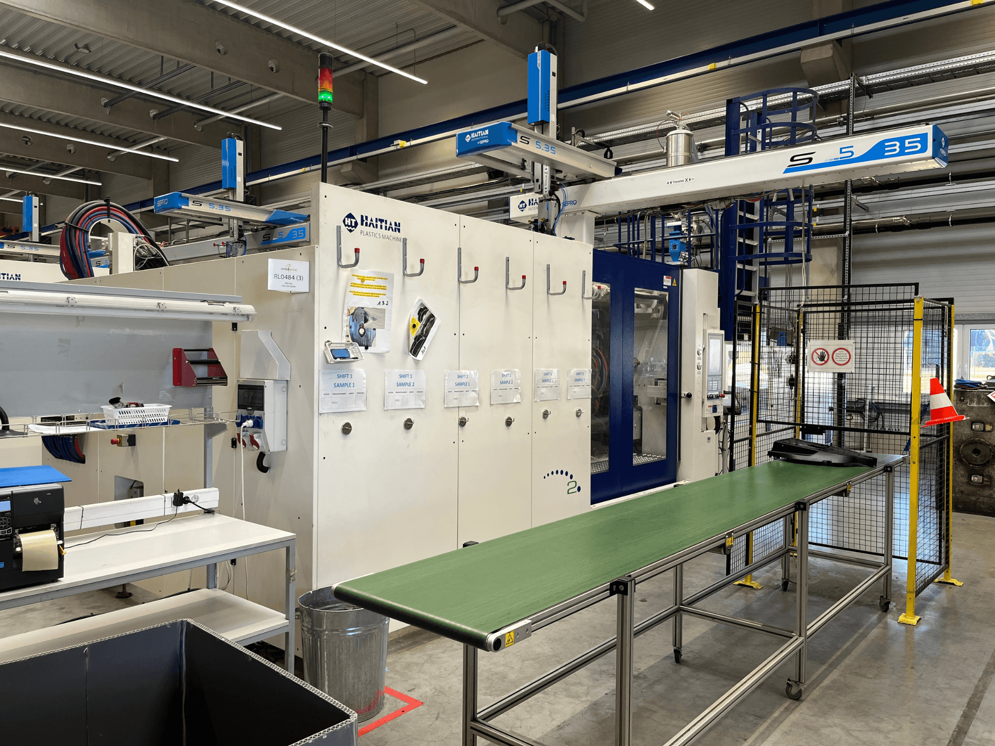 Haiti plastmasas mašīna S5-35 darbnīcā; no priekšpuses redzams zaļš konveijera galds, drošības barjeras un vadības panelis.