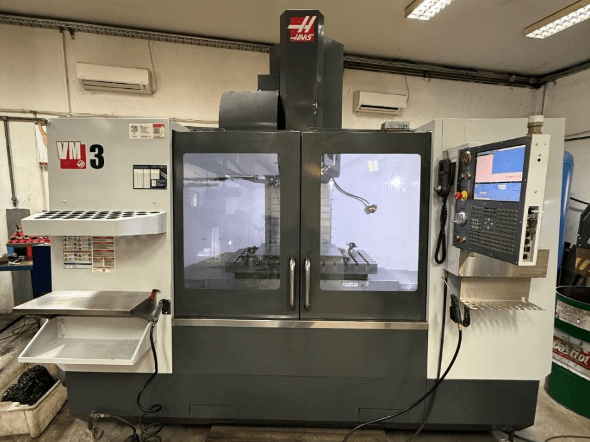 Haas VM-3 vertikālais apstrādes centrs, skats no priekšpuses, kurā redzams liels vadības panelis un darba zona ar instrumentiem.