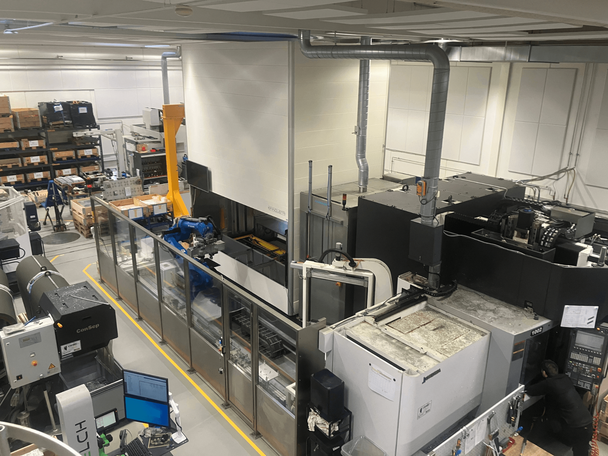 SSI SCHAFER Logimat automatizācijas sistēma noliktavā, skats no priekšpuses; ar robotizētu roku, glabāšanas plauktiem un iekārtām.