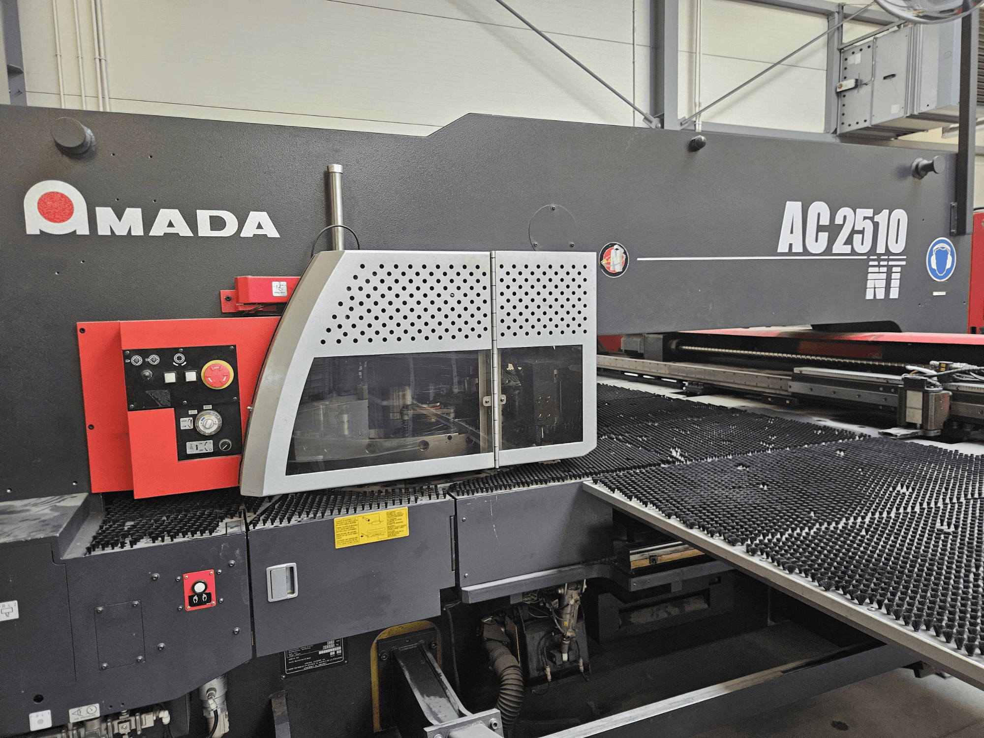 Amada AC2510 NT lāzera griešanas iekārta, skats no priekšpuses, ar vadības paneli ar pogām, pogām un drošības vāku.