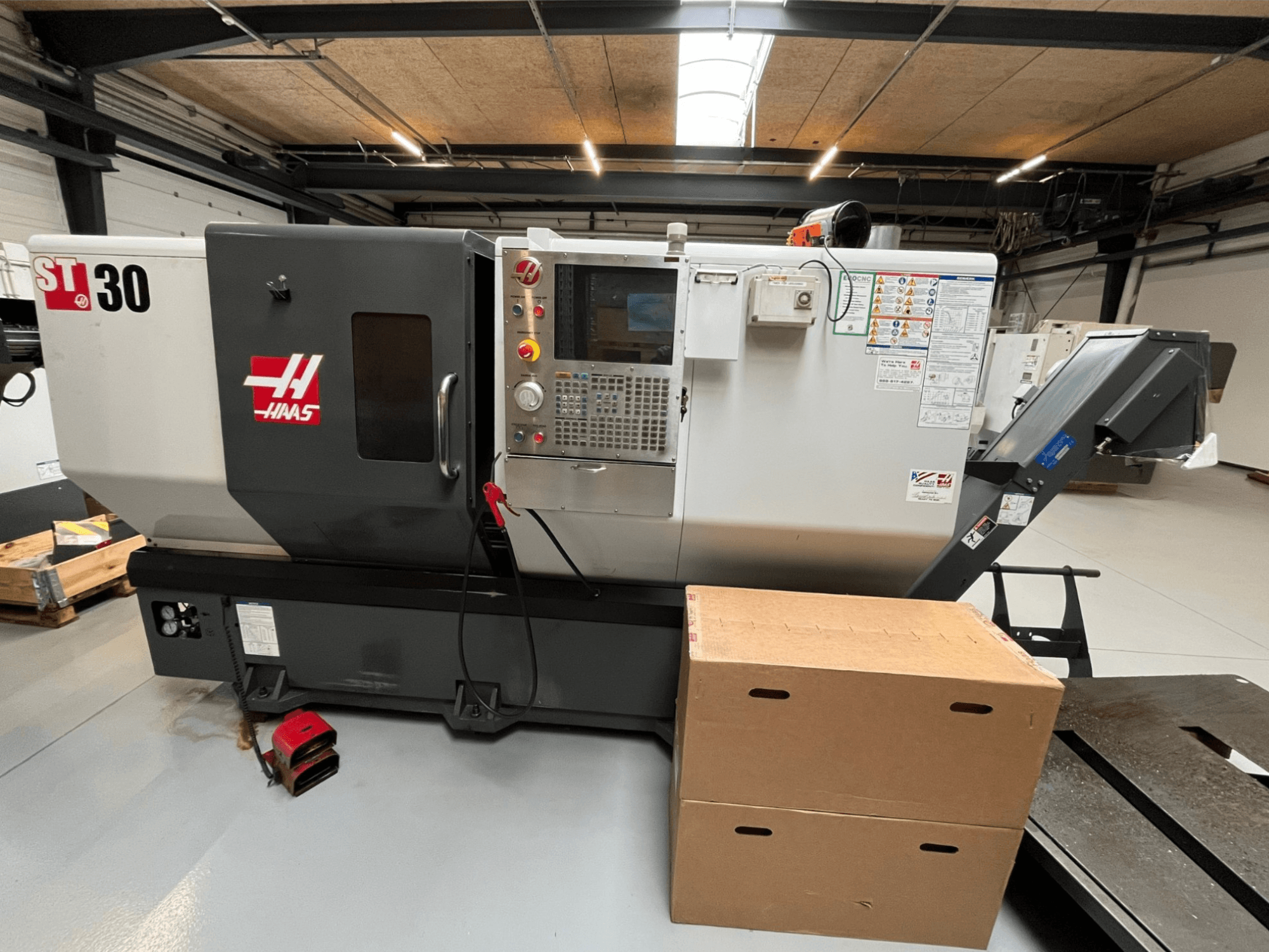 Haas ST-30 CNC virpa, skats no priekšpuses, vadības panelis ar pogām, avārijas apstāšanās un divas kartona kastes.