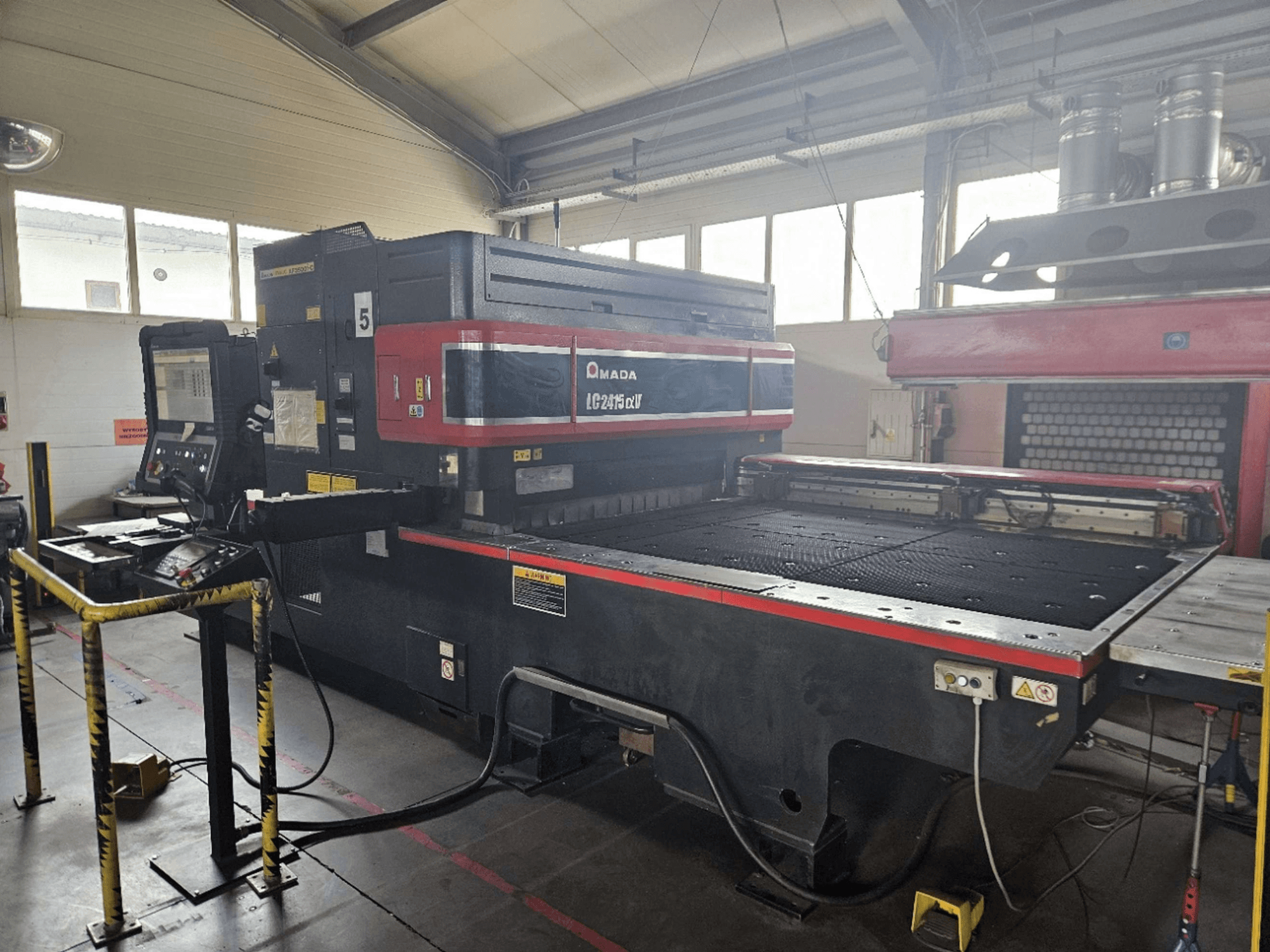 AMADA LC2415 Alpha 5 CO₂ lāzergriešanas iekārta, skats no priekšpuses, kurā redzams vadības panelis un griešanas galds ar drošības barjerām.