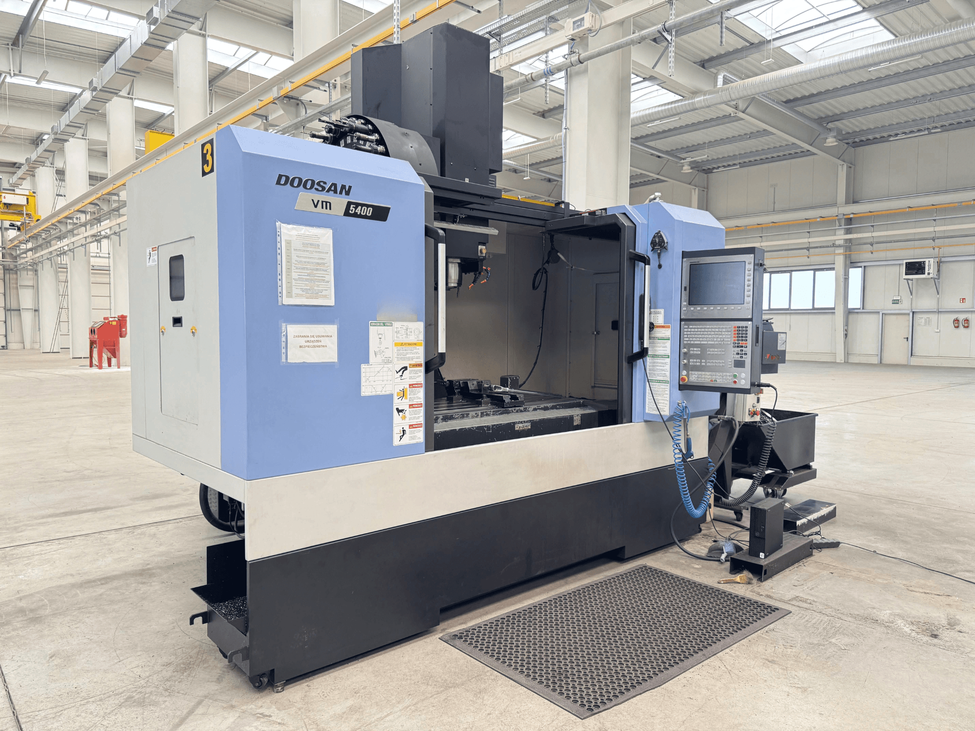 DOOSAN VM 5400 vertikālais apstrādes centrs, priekšējais skats, kurā redzams vadības panelis un darba zona plašā rūpnieciskā vidē.