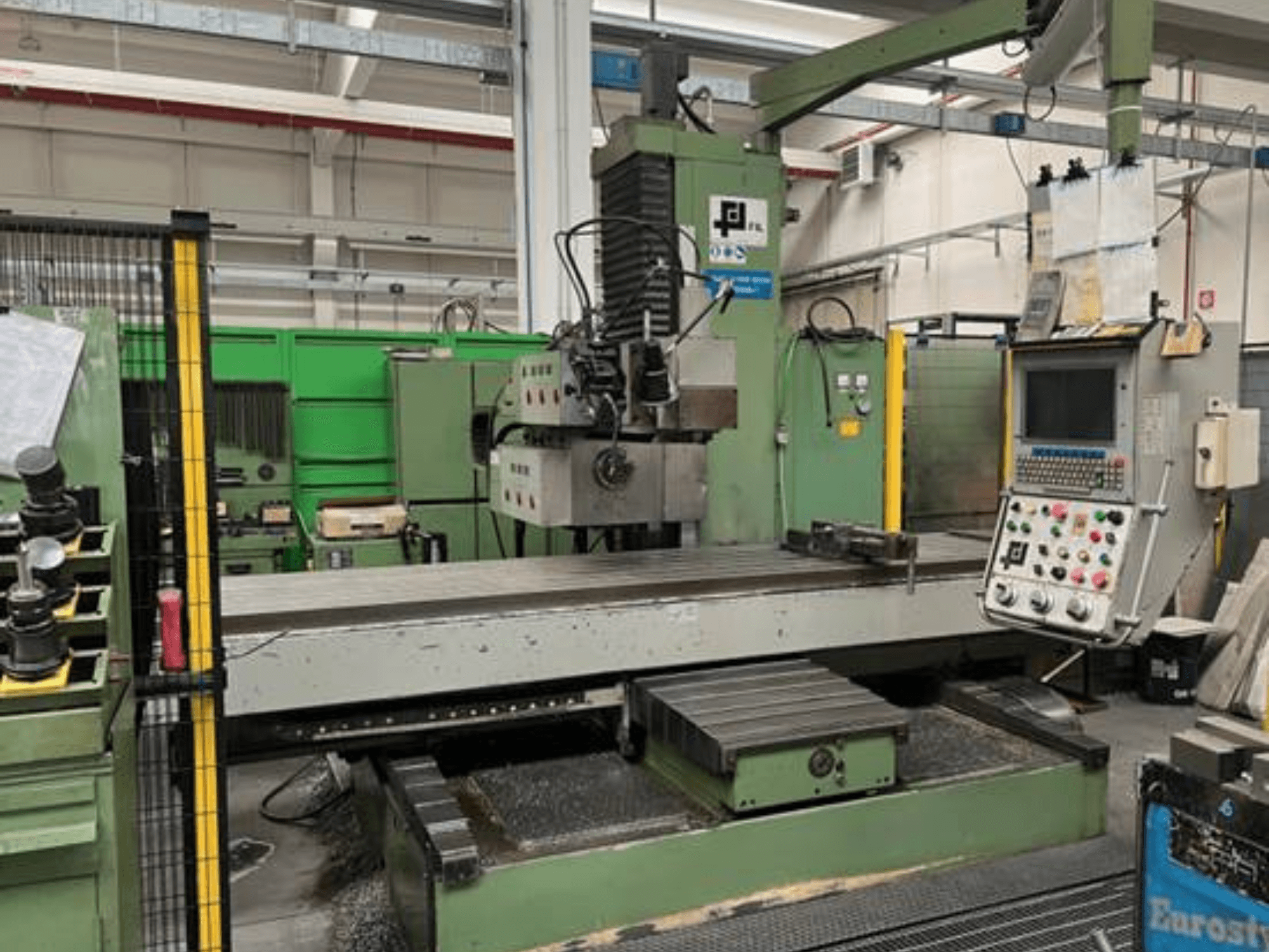 Rūpnieciskā frēzmašīna no JFL, priekšējais skats ar vadības paneli, lielu plakanu galdu un metāla skaidām uz grīdas.