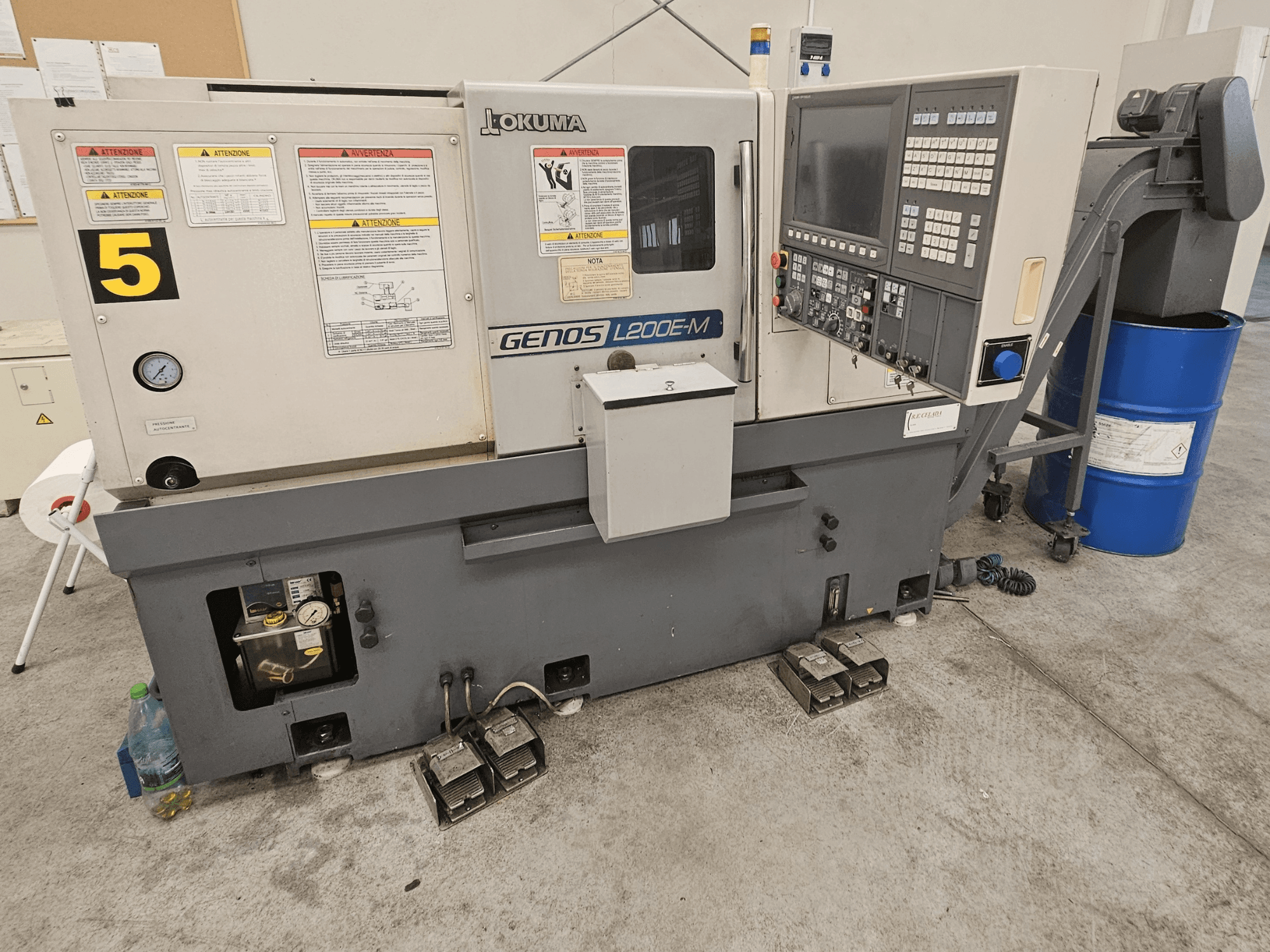 Okuma GENOS L200E-M horizontālās virpošanas darbgalds priekšpusē ar vadības paneli, etiķetēm un dzesēšanas šķidruma mucu.