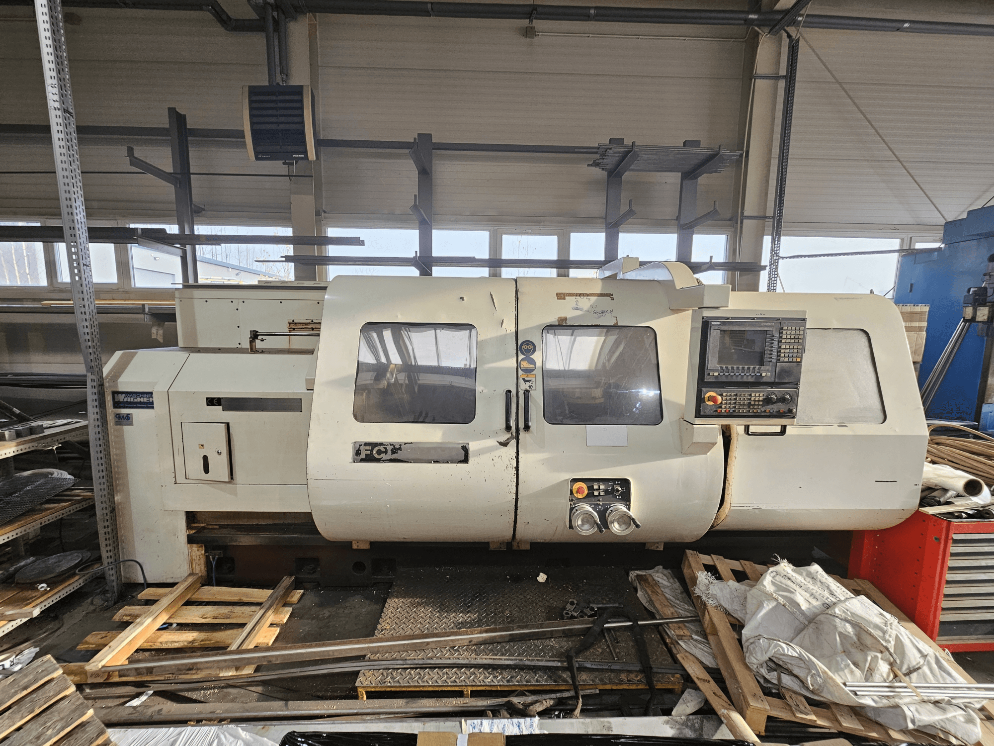 Wabeco CNC virpa, skats no sāniem, ar vadības paneli, logiem un darba zonu; iekārtas uz koka paliktņiem darbnīcā.