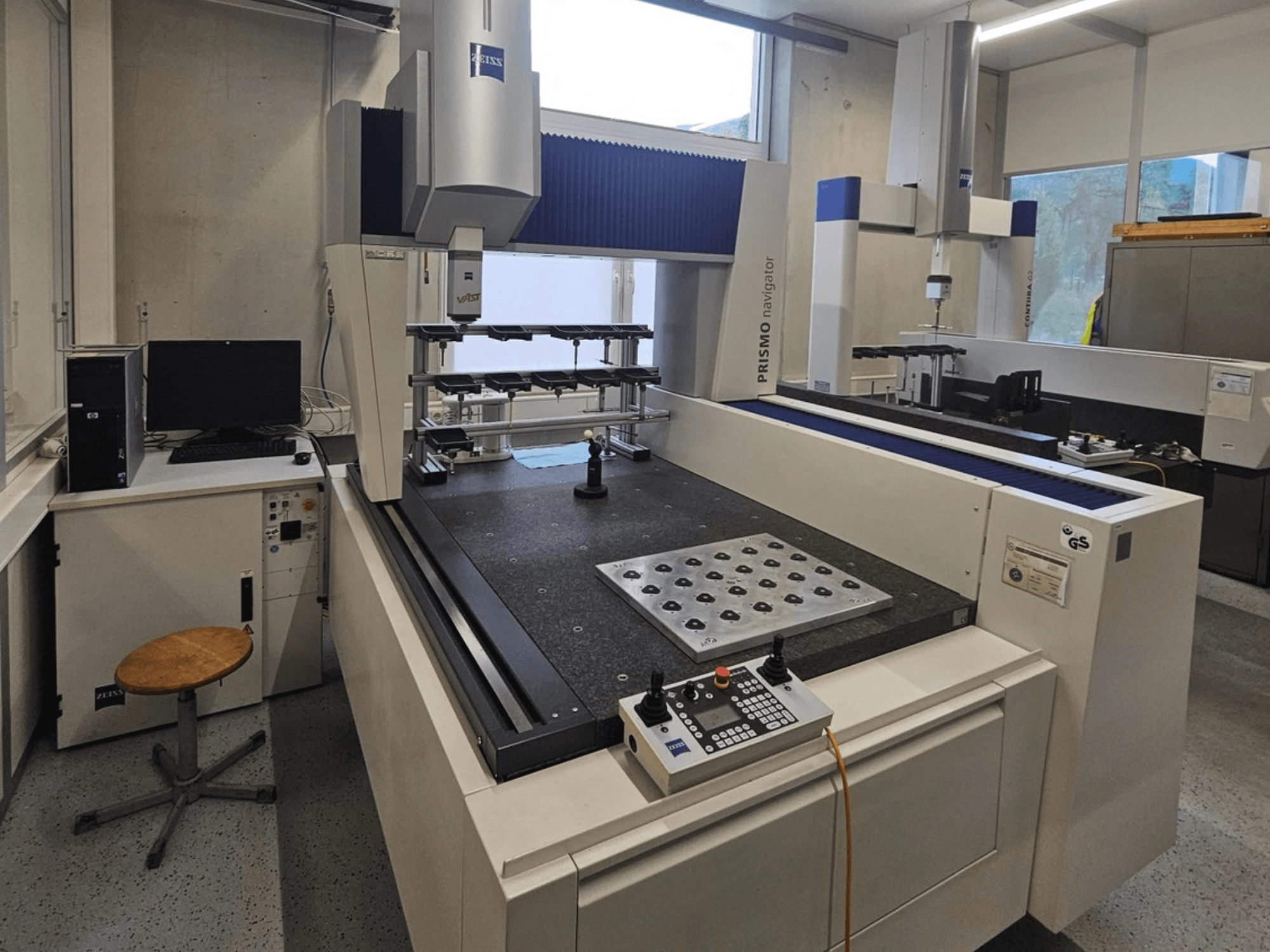 ZEISS Prismo Navigator 7 CMM laboratorijā — skats no priekšpuses, kurā redzams vadības panelis, darba galds un monitors.