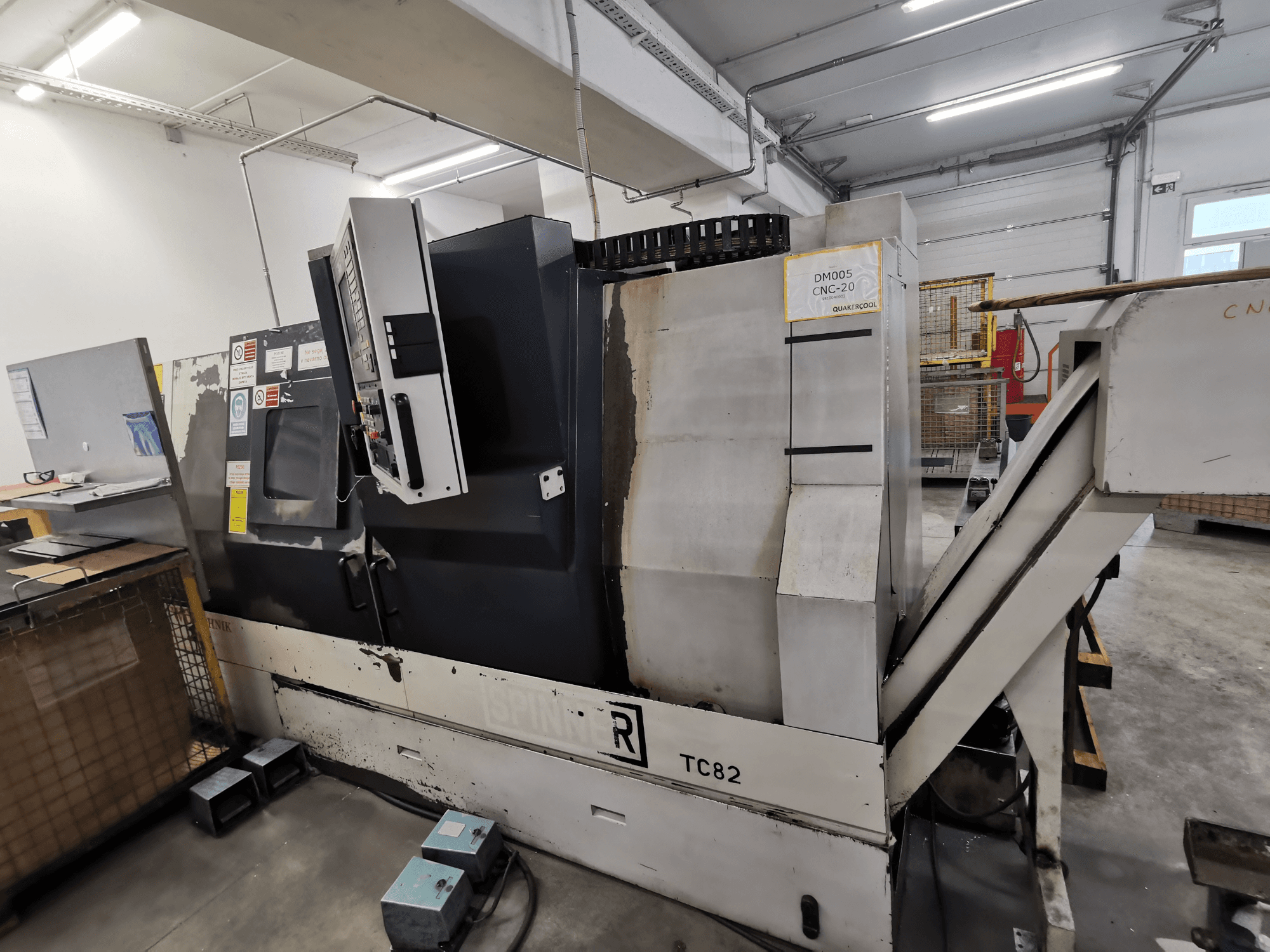 CNC mašīna, zīmols Spinner, modelis TC82, skats no sāniem, kurā redzams vadības panelis un lūžņu izvešanas tekne darbnīcā.