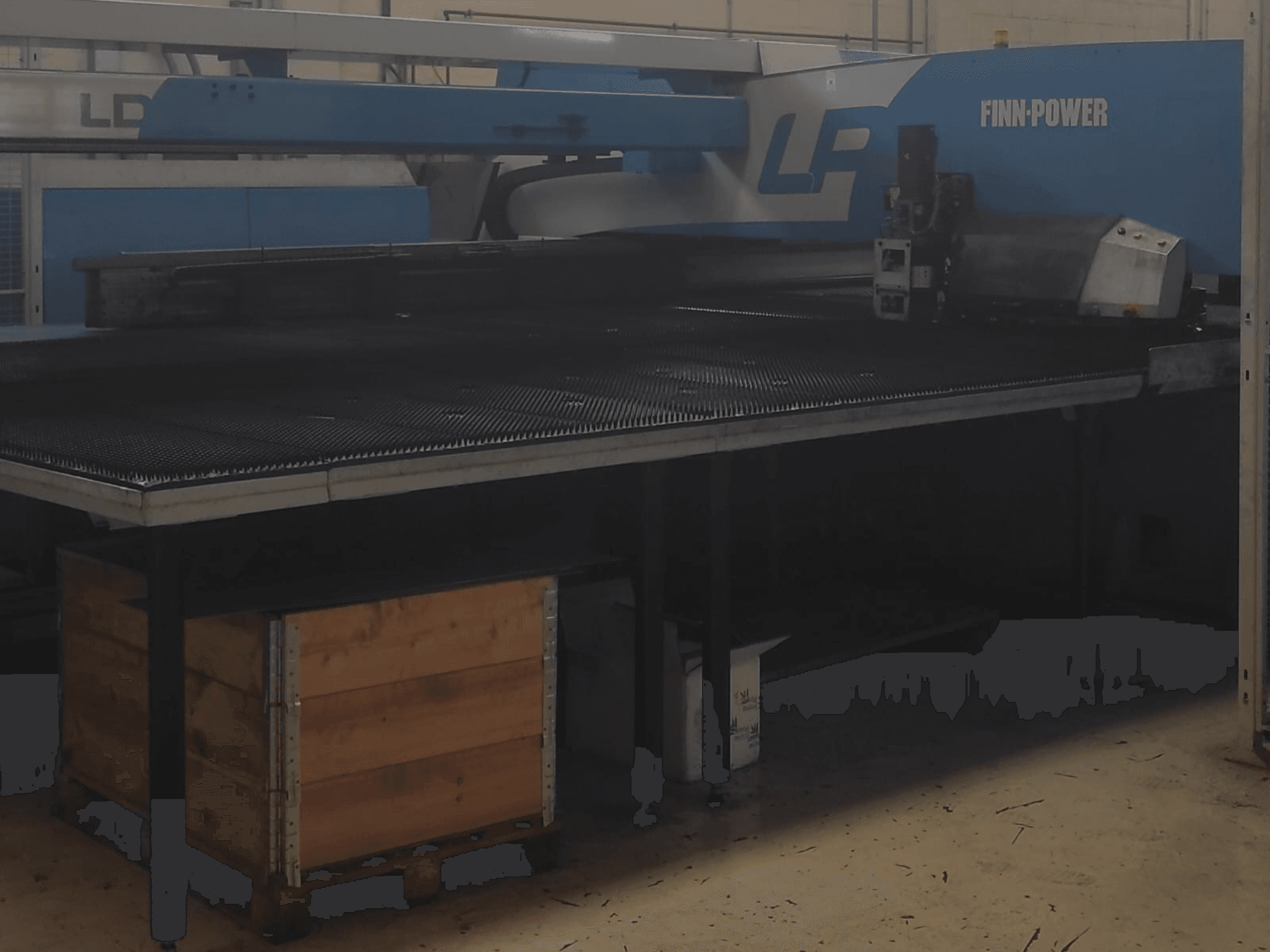 Finn-Power LP griešanas iekārta rūpnīcā, skats no priekšpuses ar teksturētu metāla galdu un koka kasti zem tā.