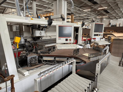 IMA precīzijas kokapstrādes darbgalds ar diviem monitoriem un vadības paneļiem, priekšējais skats, kurā redzams konveijers un instrumentu zona.