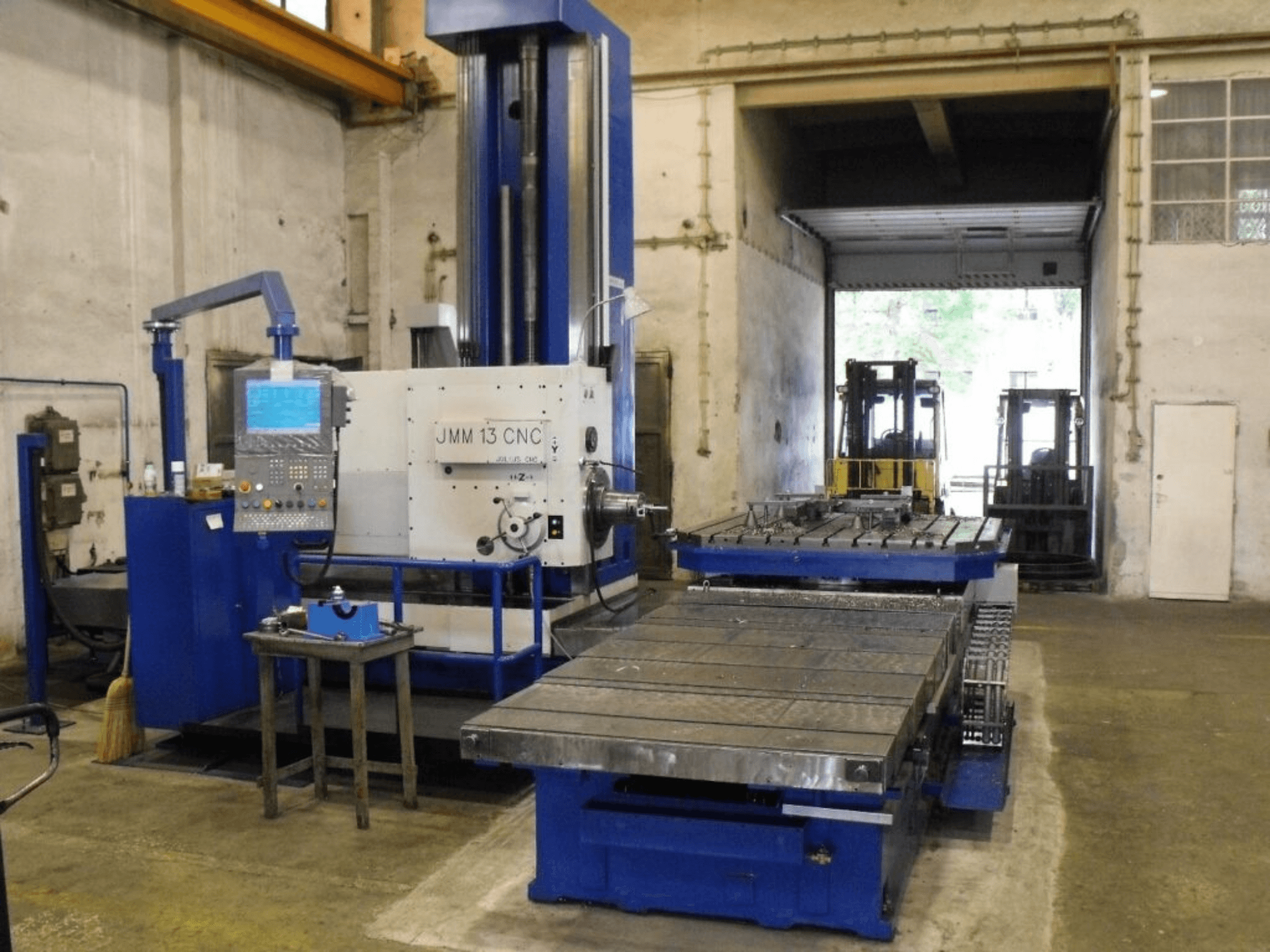 JMM 13 CNC frēzmašīna darbnīcā, skats no priekšpuses, ar vadības paneli, lielu darba galdu un rūpniecisku fonu.