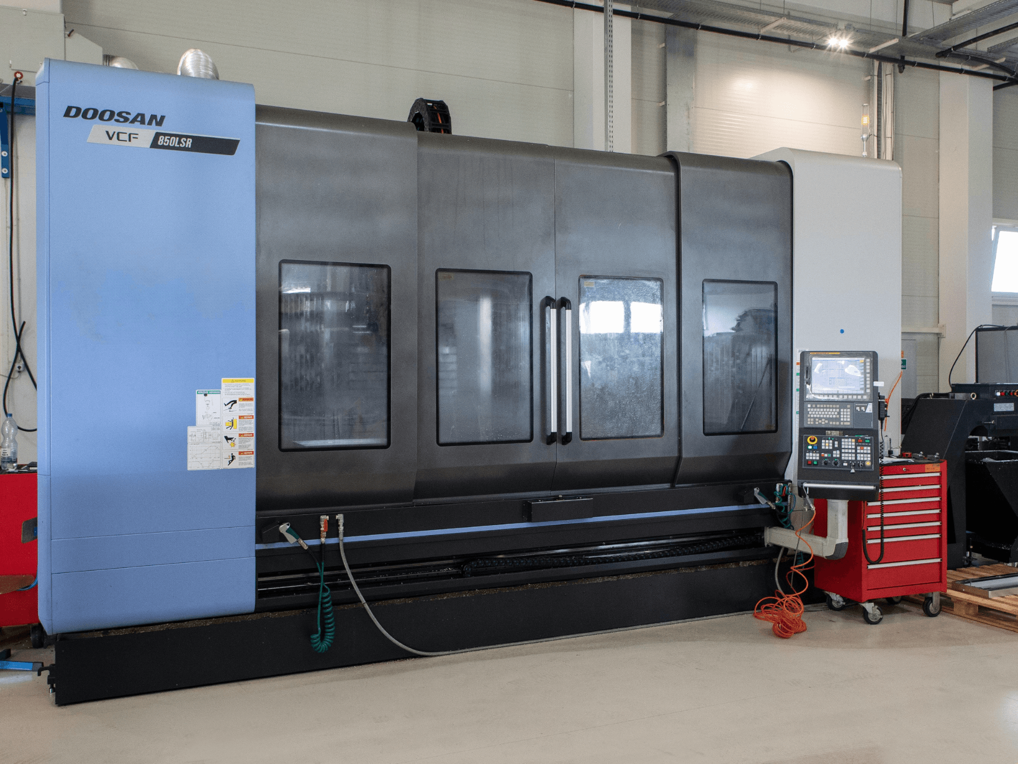 DOOSAN VCF850 frēzēšanas-virpošanas centrs, skats no priekšpuses; aprīkots ar lieliem dubultlogiem, vadības paneli un instrumentu glabāšanas nodalījumu. Ideāli piemērots metālapstrādei.