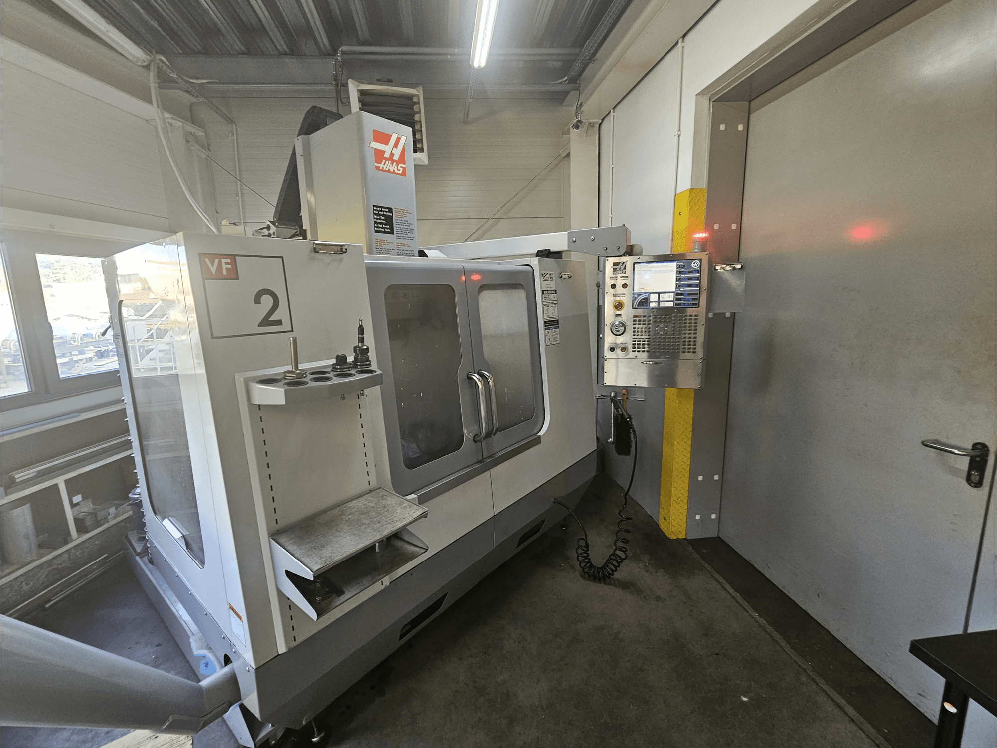 Haas VF-2 CNC frēzmašīna, skats no priekšpuses, kurā redzams vadības panelis, instrumentu turētāji un caurspīdīgs logs uz darba vietu.