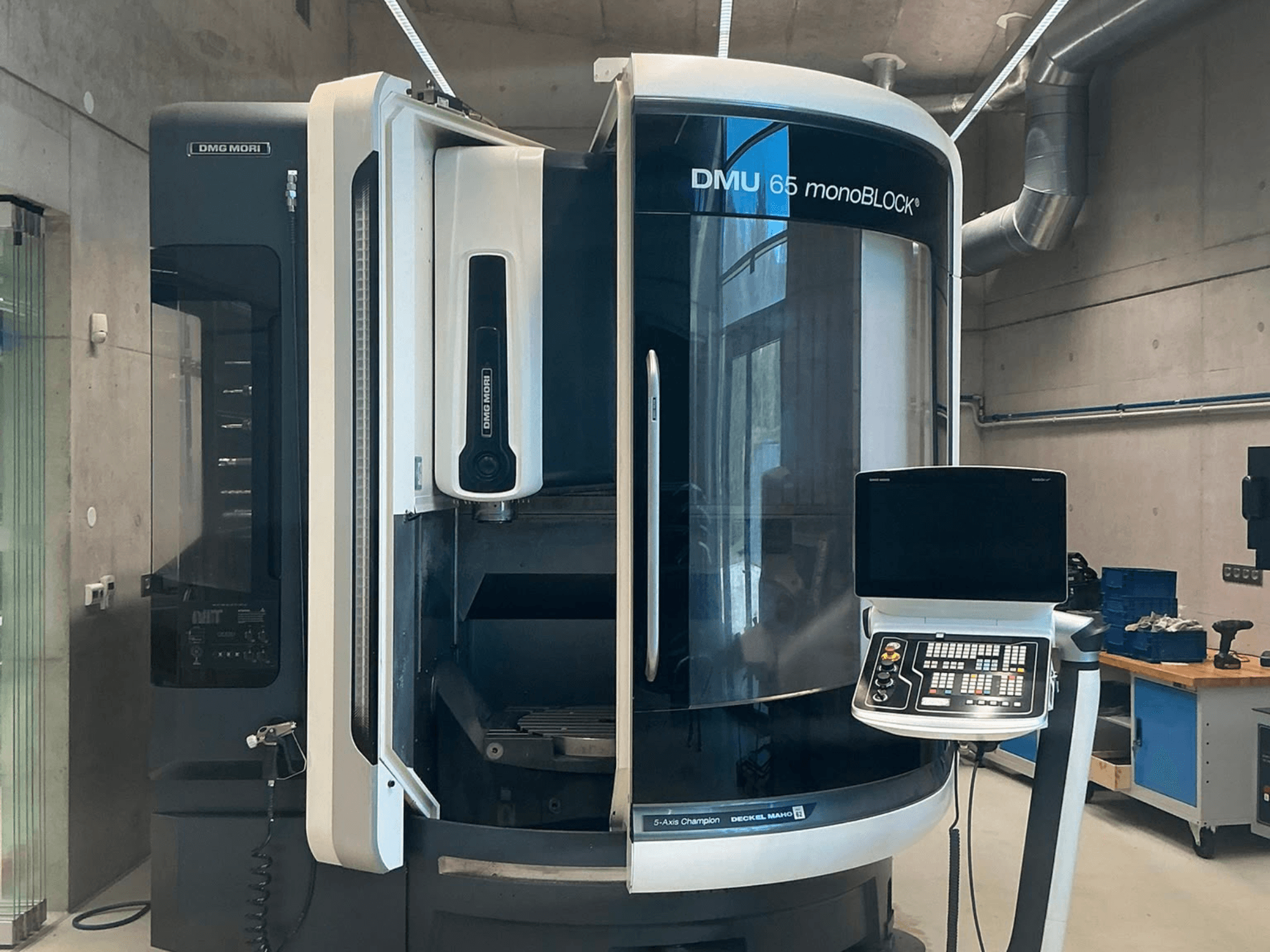 DMG MORI DMU 65 monoBLOCK vertikālais apstrādes centrs, skats no priekšpuses, kurā redzams vadības panelis un mašīnas detaļas darbnīcas vidē.