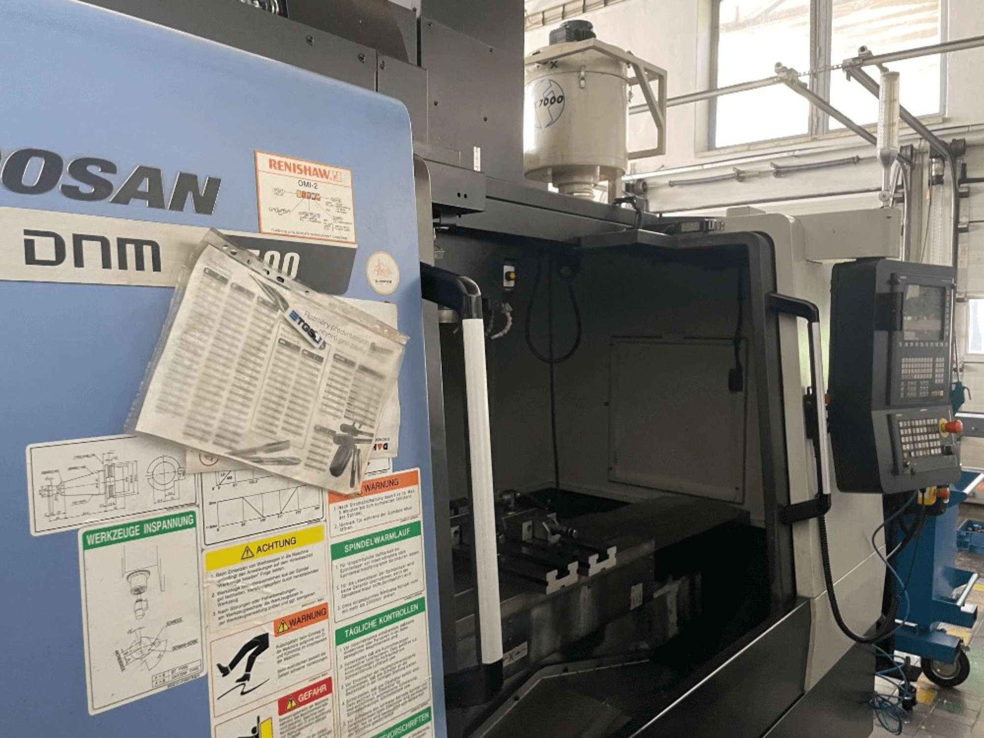 DOOSAN DNM 5700 vertikālais apstrādes centrs, skats no priekšpuses, redzams vadības panelis, drošības marķējums un mašīnas detaļas.