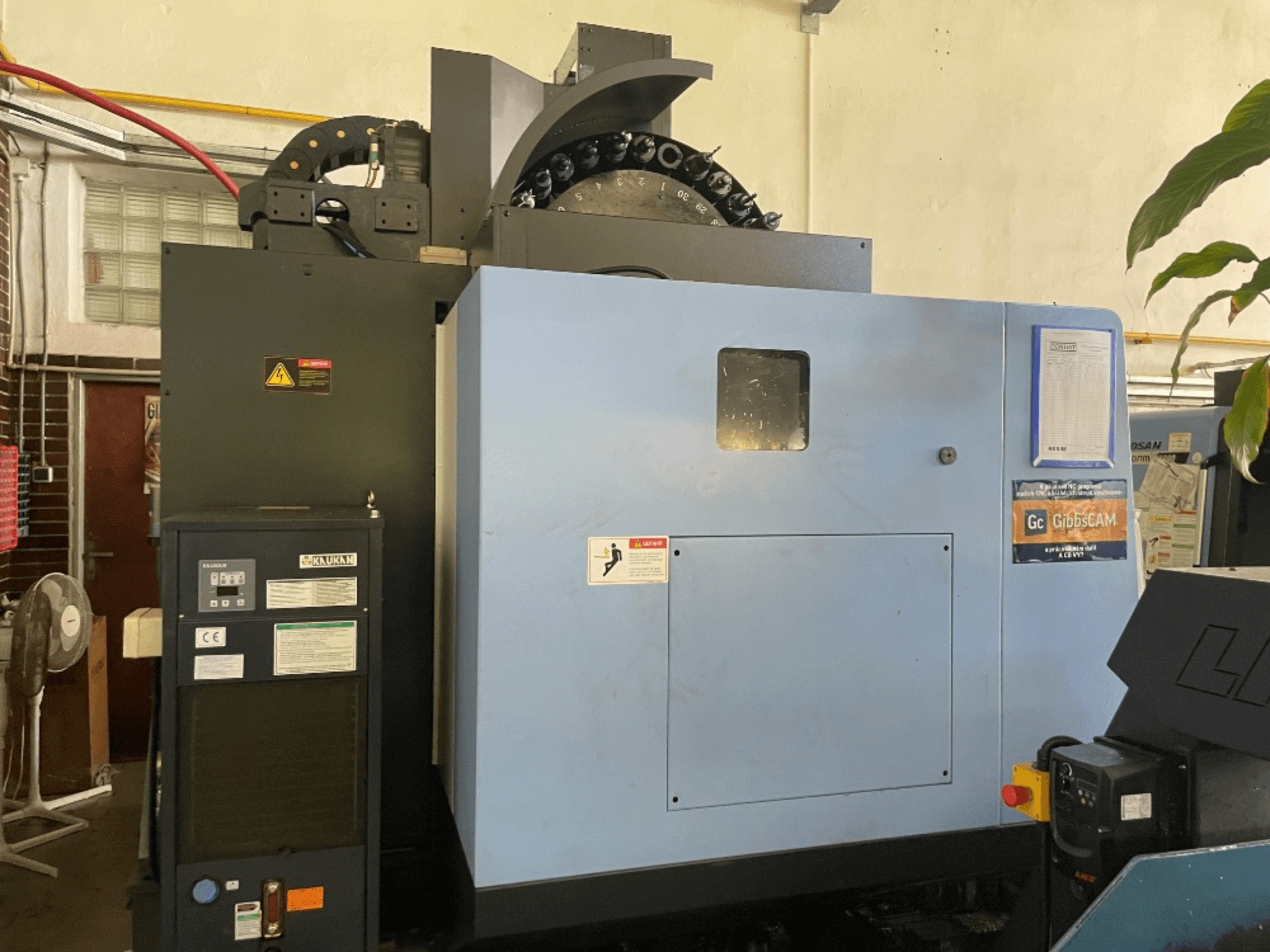 Vertikālā apstrādes centra DOOSAN DNM 5700 priekšējais skats, kurā redzams vadības panelis, drošības marķējums un mašīnas funkcijas.