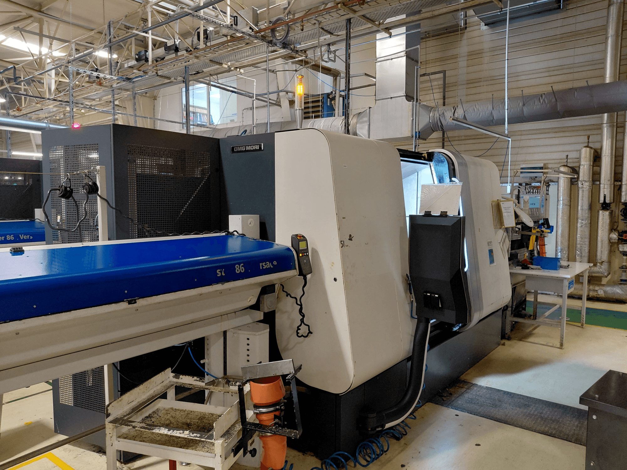 DMG MORI Sprint 65 lineārā daudzvārpstu virpošanas mašīna, skats no sāniem ar darbnīcā redzamu vadības paneli un konveijeru.