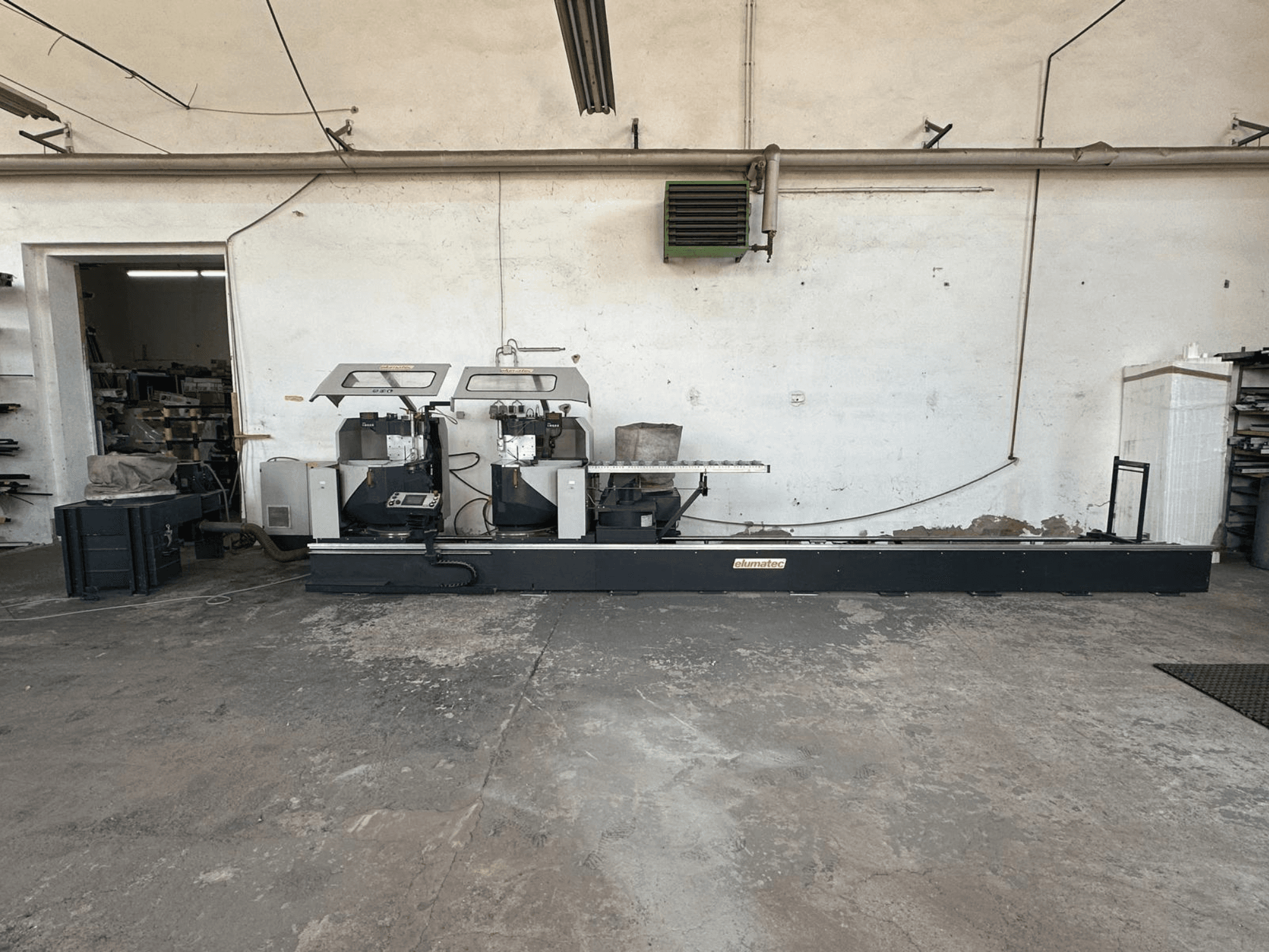 Elumatec DG 244 cirkulārais metāla zāģis darbnīcā, skats no sāniem, kurā redzami divi zāģi un vadības ierīces uz izturīga pamatnes.