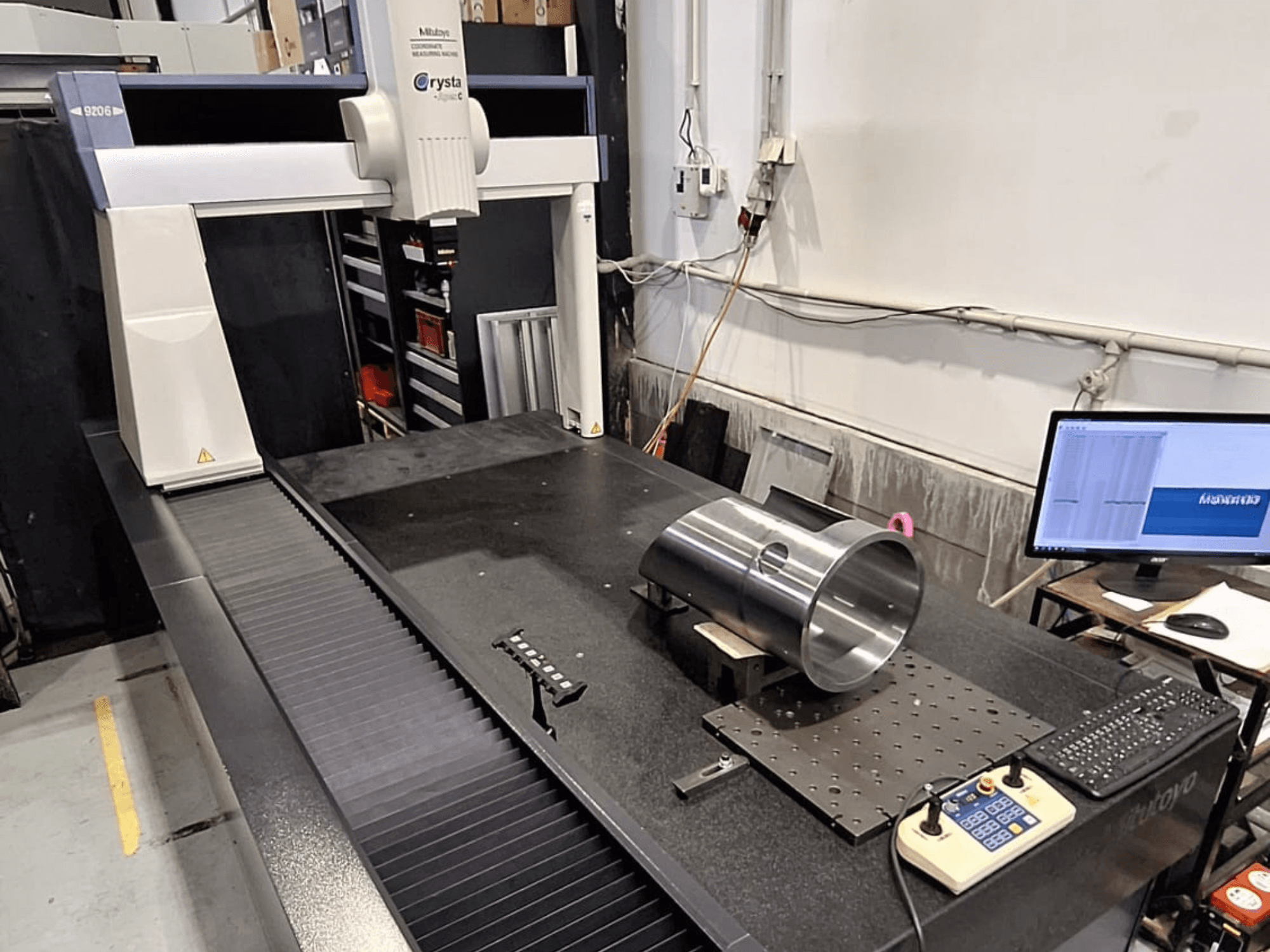 Mitutoyo Crysta-Apex C9206 CMM priekšpusē, mēra cilindrisku metāla detaļu uz melna granīta virsmas plāksnes.