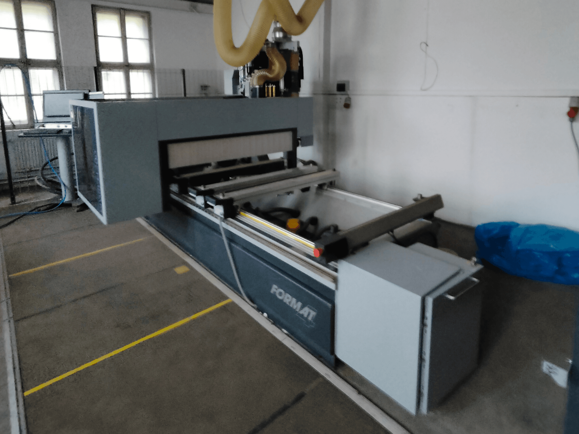 Format CNC mašīnas priekšējais skats ar pelēku korpusu, vadotnēm un redzamu vadības paneli.