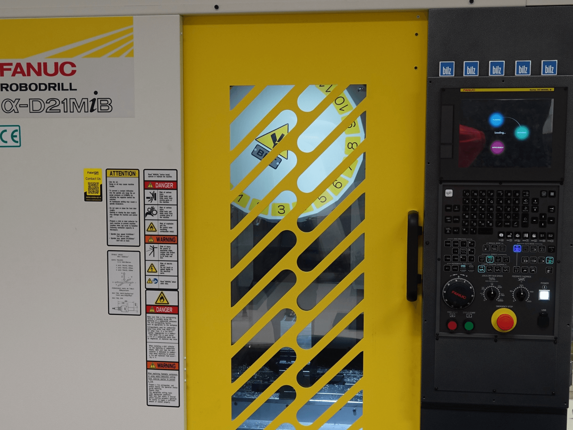 FANUC α-D21MiB vertikālā apstrādes centra skats no priekšpuses, kurā redzami drošības brīdinājumi, vadības panelis un dzeltenās drošības durvis.
