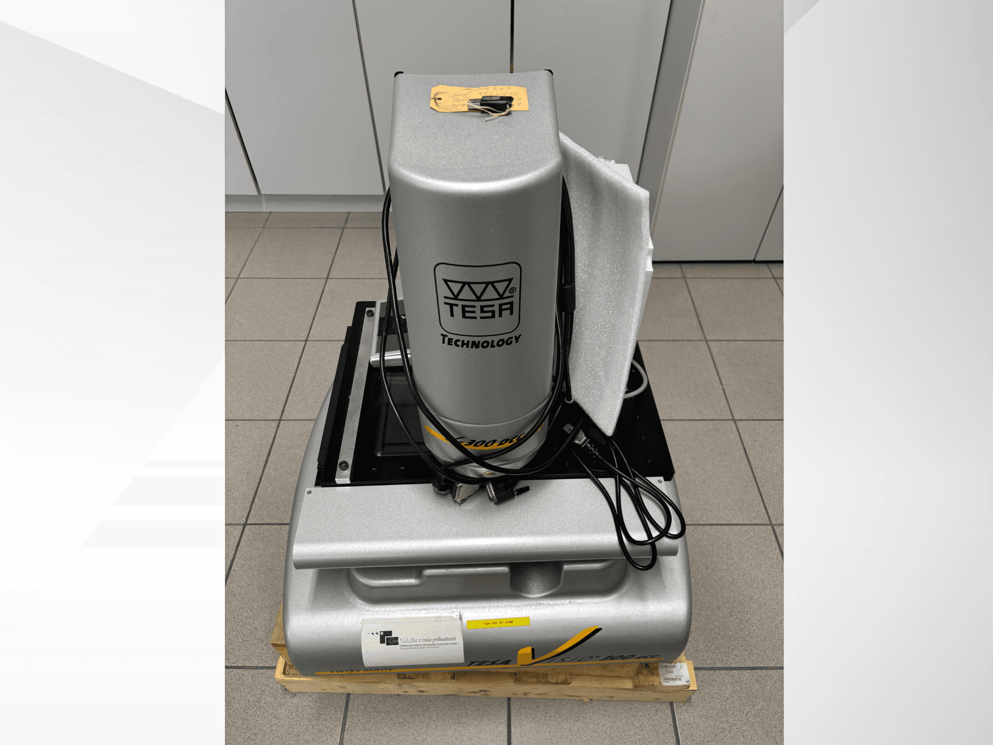 TESA Visio 300 DCC optiskā mērīšanas iekārta priekšpusē ar kabeļiem, uz koka paliktņa un redzamu aizsargapvalku.