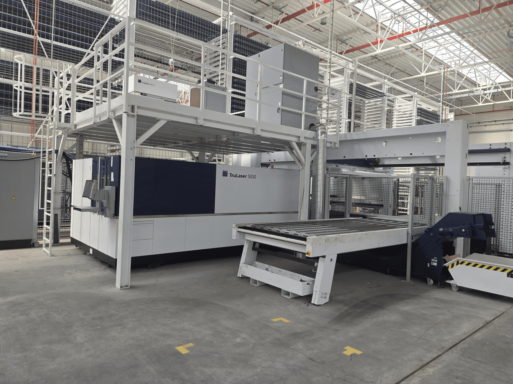 TRUMPF TruLaser 5030 fiber L76 FMC iekārta darbnīcā, skats no priekšpuses, kurā redzama griešanas zona un iekraušanas/izkraušanas mehānisms.