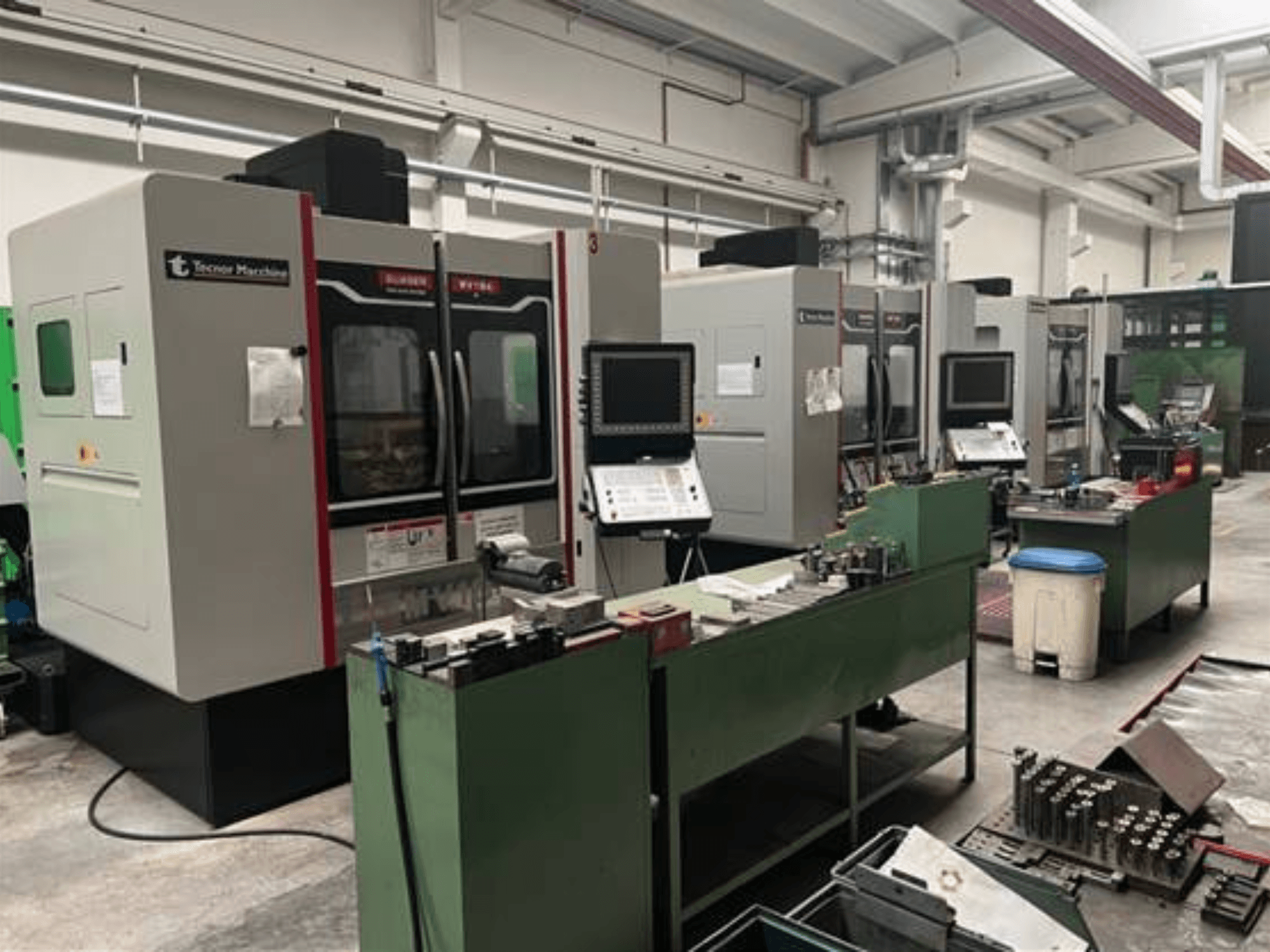 Četras Tecnoer Marchine CNC mašīnas darbnīcā, skats no priekšpuses, ar instrumentiem un detaļām uz zaļiem darbgaldiem.