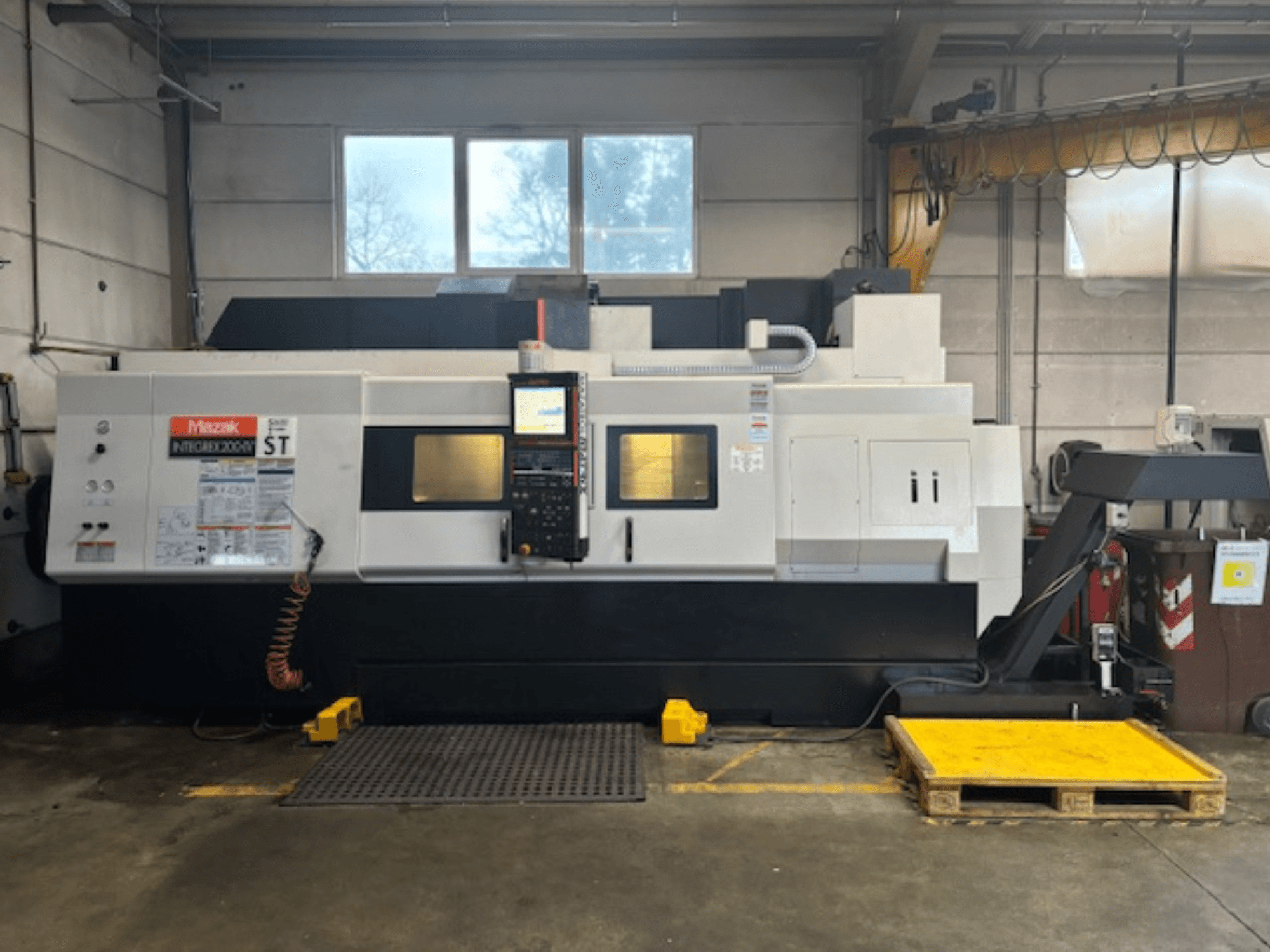 Mazak Integrex 200 SY ST, skats no sāniem, kurā redzams vadības panelis, darbības displeji un dzesēšanas šķidruma līnijas darbnīcā.