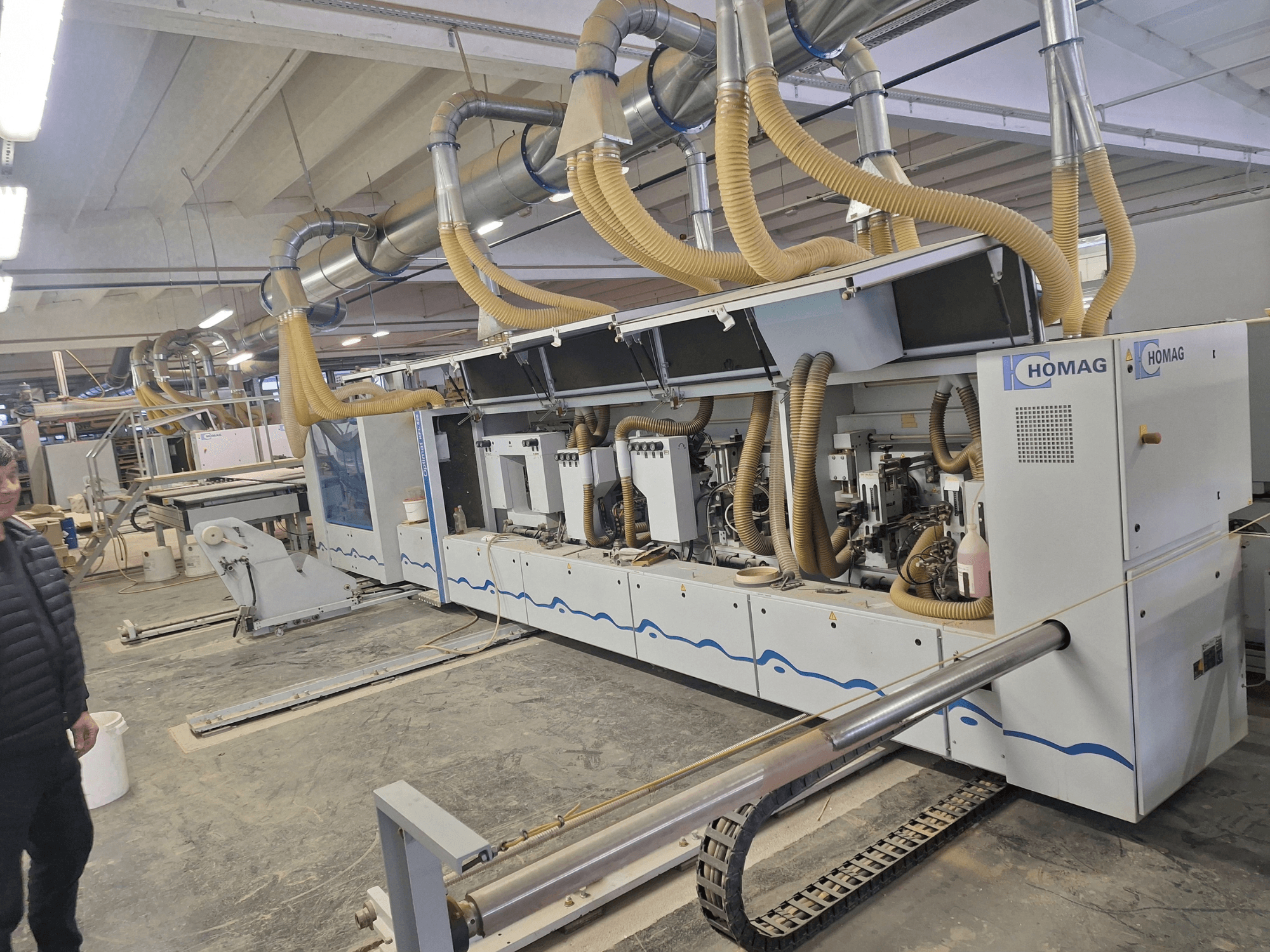 HOMAG kokapstrādes mašīna rūpnīcā, priekšējais skats, kurā redzami vairāki vārpsti un putekļu nosūces sistēma.