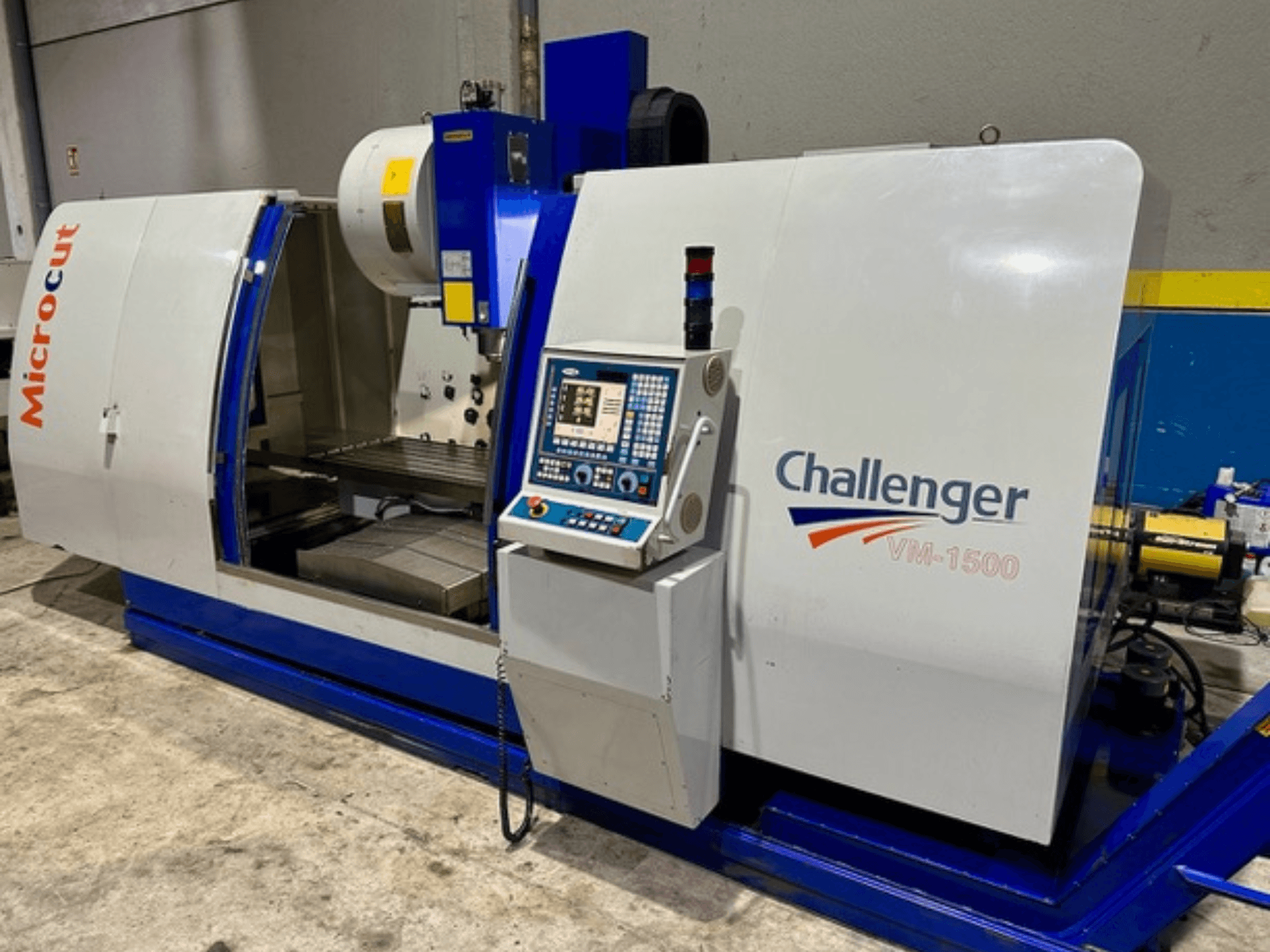 Microcut Challenger VM-1500 CNC darbgalds, skats no priekšpuses, ar vadības paneli, darba vietu un zilu pamatni.